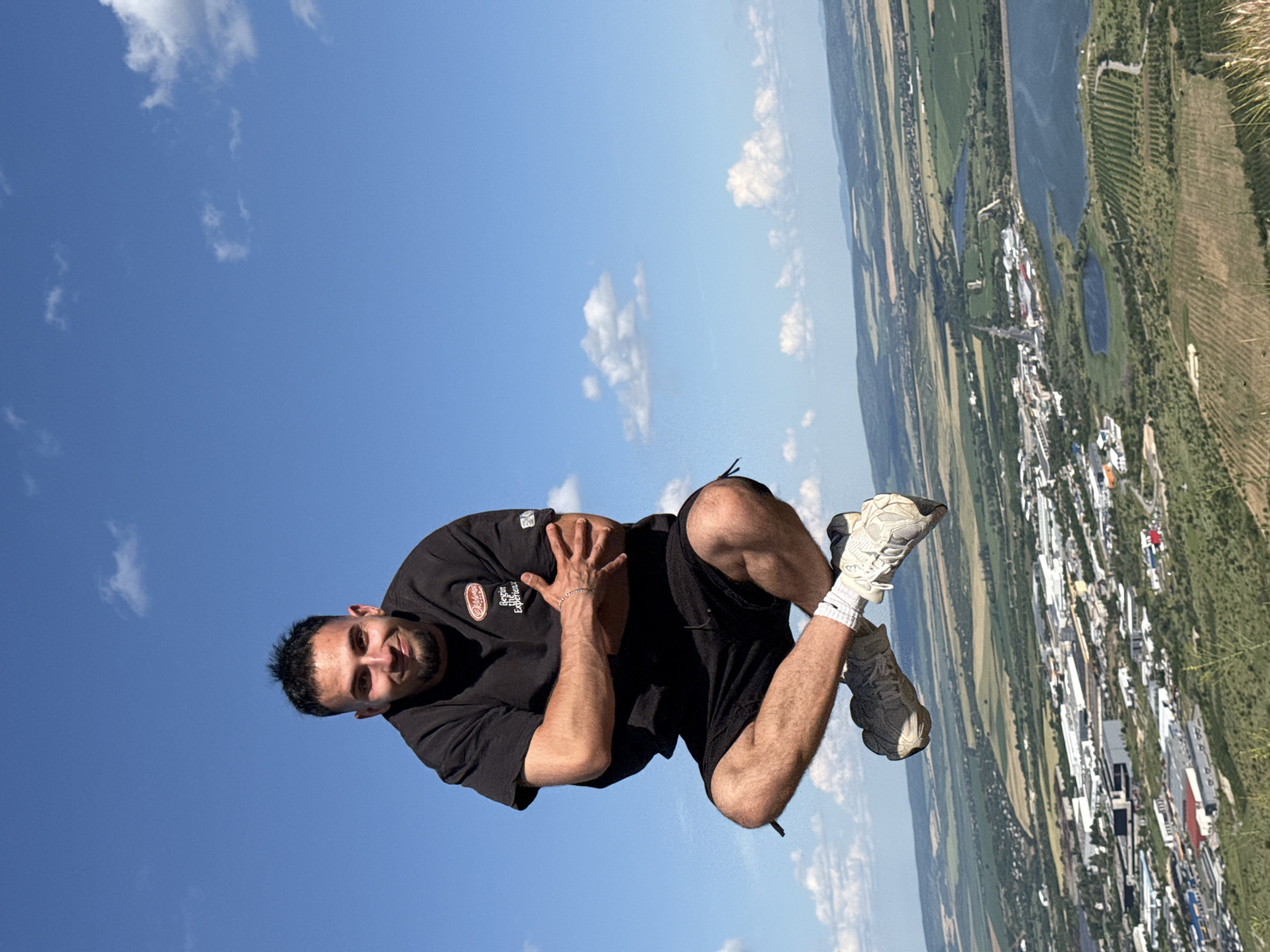 A man in a black t-shirt and black shorts is captured mid-air in a levitating pose, appearing to be jumping from a hilltop overlooking the city of Shumen, Bulgaria. He has his arms crossed and is smiling at the camera. His legs are bent, with one foot crossing over the other, and he is wearing white socks and grey sneakers. The background reveals a sprawling cityscape with buildings, roads, and bodies of water, set against a backdrop of green hills and a bright blue sky with scattered white clouds. The perspective suggests the photograph was taken from a high vantage point, likely from a significant elevation within or near the Shumen Plateau, offering a panoramic view of the city and its surroundings.