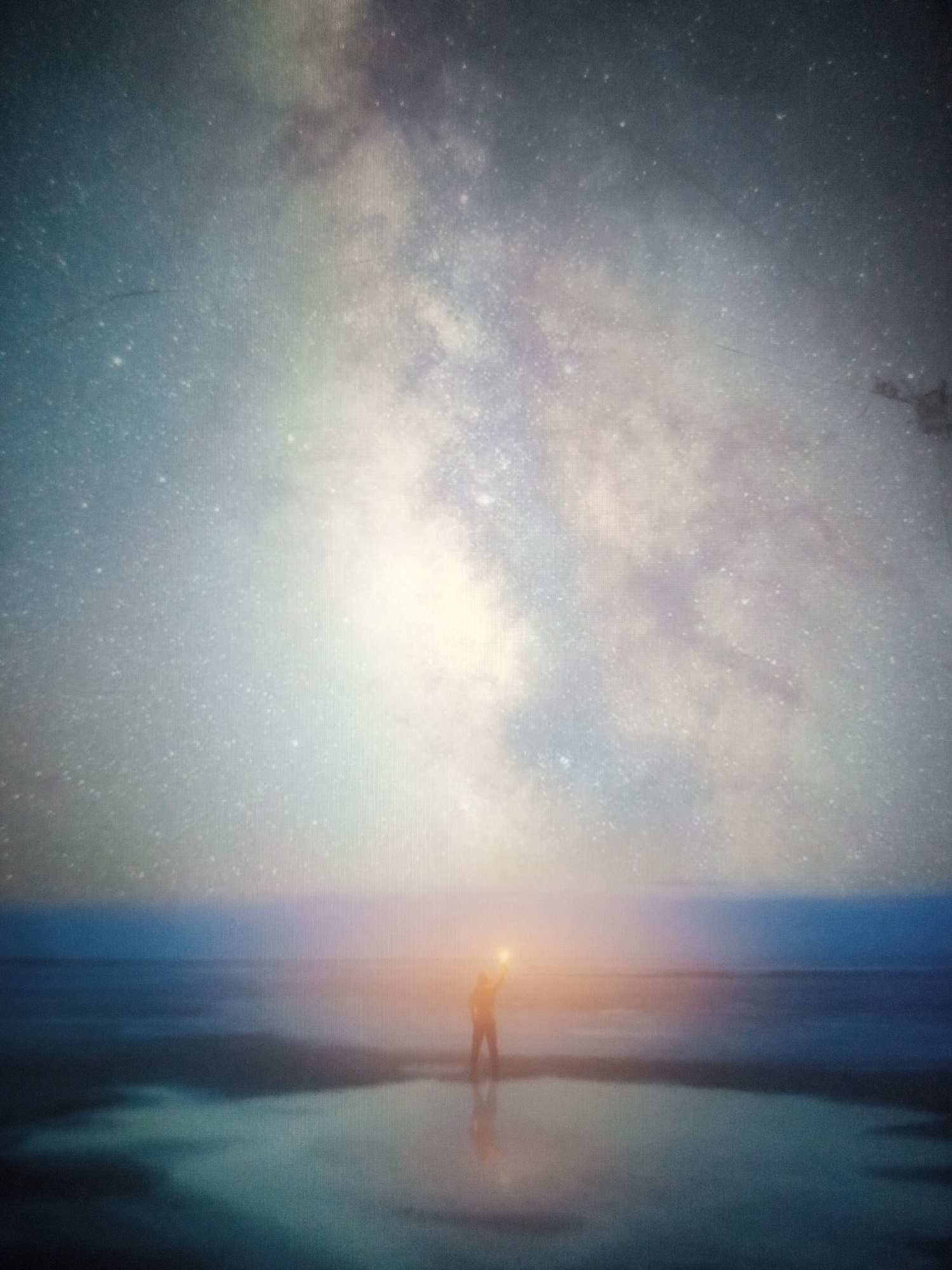 A person stands in a reflective body of water under a starry sky, holding a light aloft. The person is silhouetted against a bright light source. The water reflects the sky and the person. The sky is filled with stars and a bright band of the Milky Way. The overall scene is dreamlike and ethereal. The location is unknown.