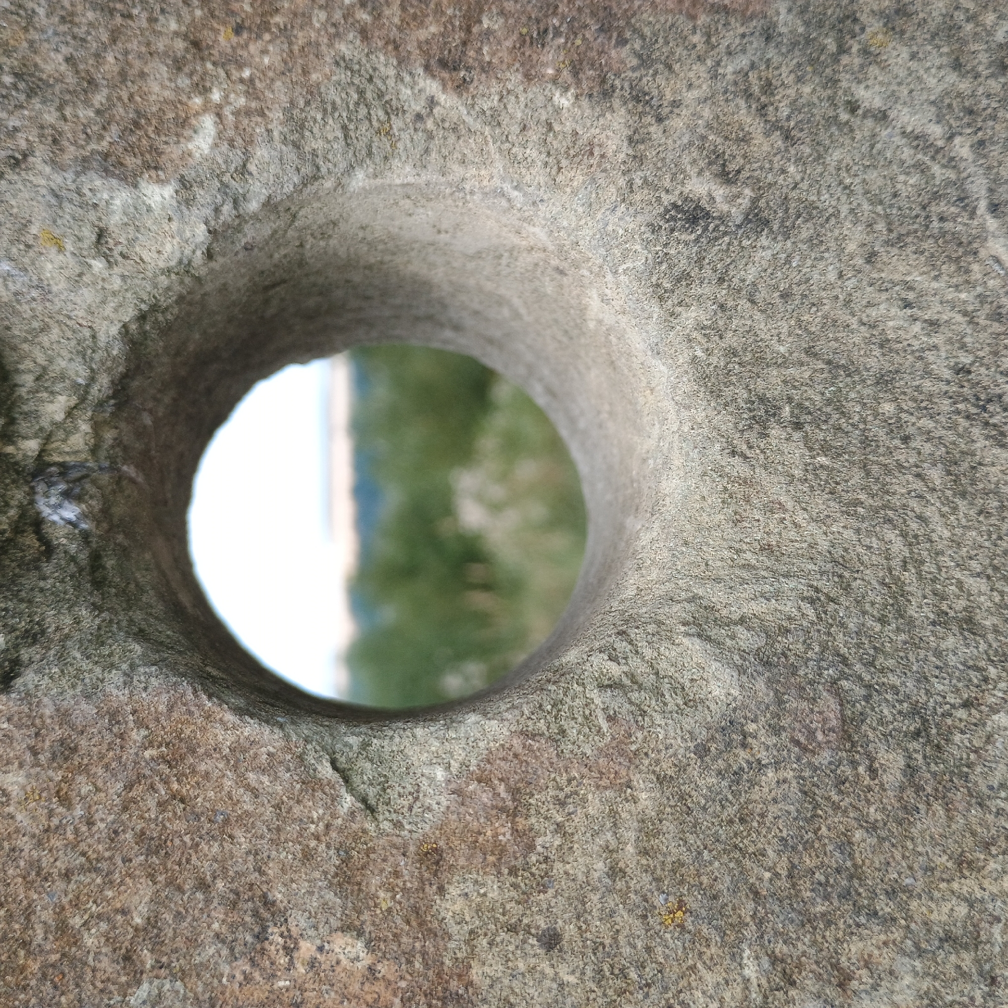 A close-up, eye-level shot focuses on a roughly circular hole carved through a large, weathered, grey stone. The stone's surface is textured with small granular particles and shows variations in color from light grey to brown and speckled dark grey. Some small yellow and white specks are visible on the stone. The hole, appearing to be a cutout or aperture, is centrally located in the frame and is slightly wider than it is tall. Through the hole, a blurred, distant landscape is visible. This landscape consists of soft green foliage, possibly trees and bushes, with a pale, hazy sky above. The scene appears to be outdoors in a natural setting. The location is identified as Möhnesee, Germany. No people or animals are present in the image.