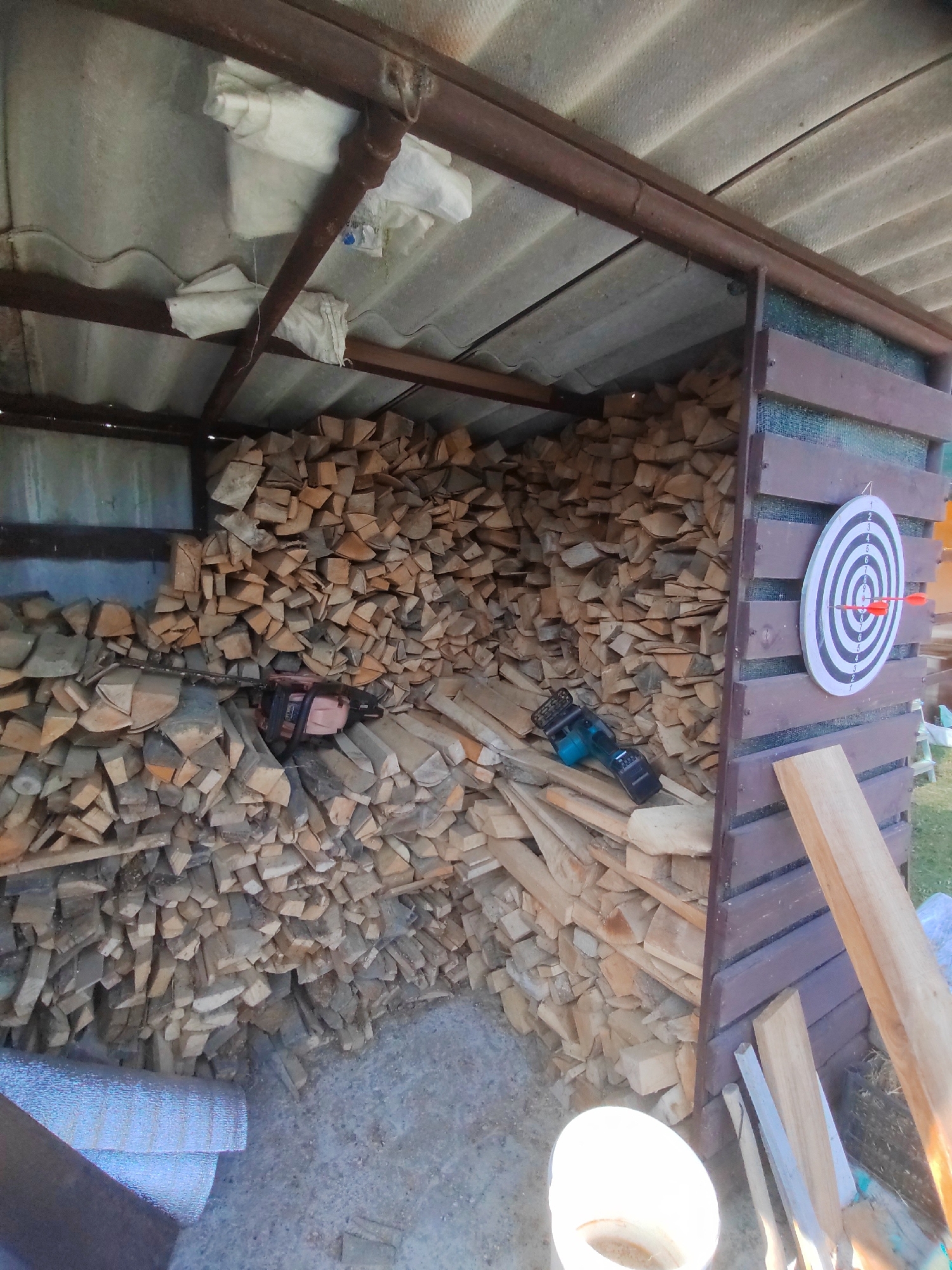 The image shows a wood storage shed filled with cut firewood. Two chainsaws are visible amongst the wood. On the exterior wall of the shed, a dartboard with two darts is mounted. A white bucket and rolls of reflective insulation material are on the ground in the foreground. The scene is set outdoors, likely in a rural or semi-rural environment. The city of Psebai, Russia, is mentioned as the location.