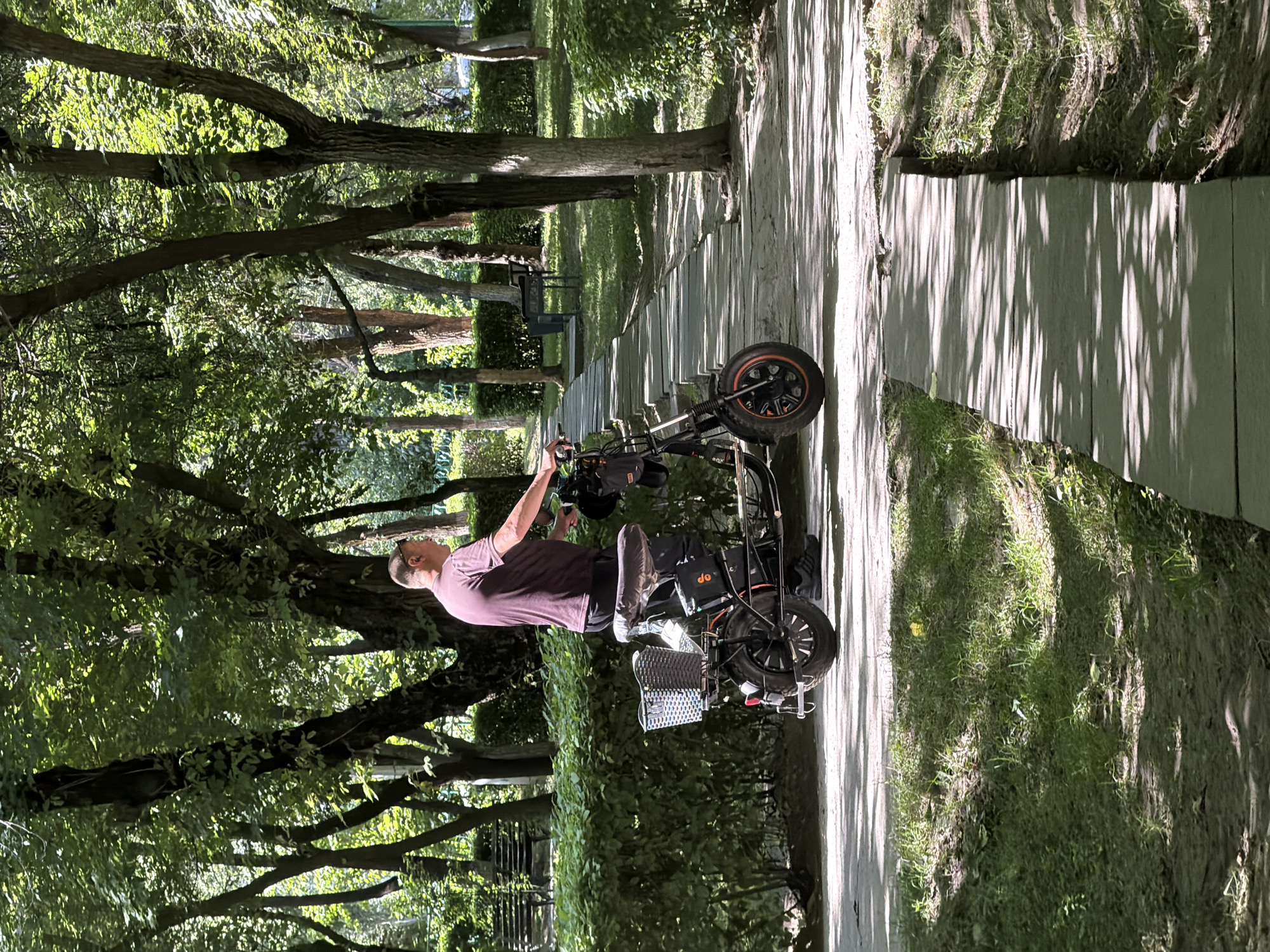 A man in a purple t-shirt and dark pants stands with his right hand on the handlebars of a black electric scooter parked on a paved path in a park. The scooter has a front basket filled with items, a black seat, and a battery pack attached to the frame. Sunlight filters through the lush green trees and foliage overhead, casting dappled shadows on the path and surrounding grassy areas. In the background, there are more trees, hedges, and park benches. The path is made of concrete slabs interspersed with grass. The scene appears to be a tranquil park setting.