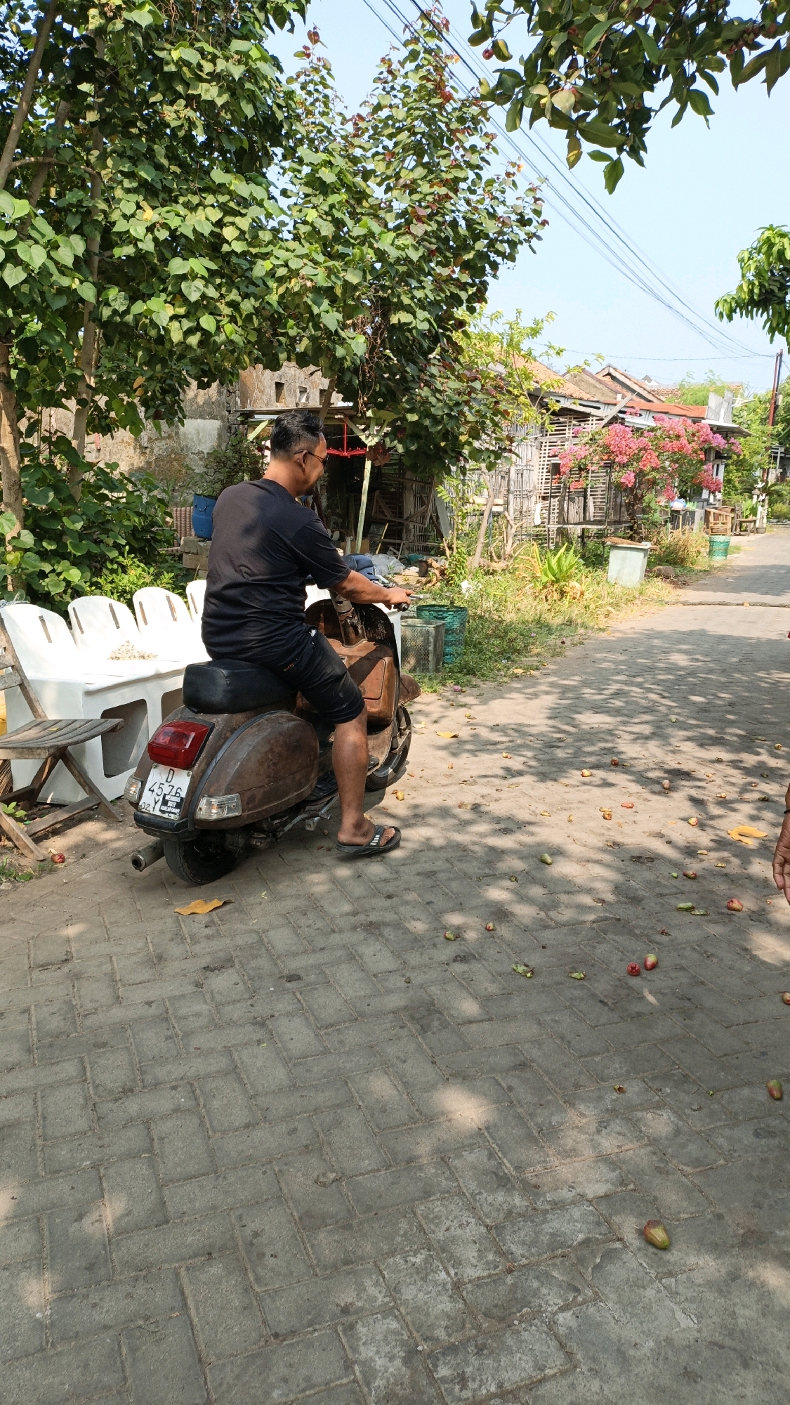 An outdoor, daytime, medium shot captures a man from the rear as he sits on a rusty, brown Vespa scooter parked on a brick-paved pathway. The man wears a black t-shirt and black shorts, with dark sandals on his feet. He has glasses on and is looking to his right, with his hands on the scooter's handlebars. The scooter has a red taillight and a license plate with the characters "D 4576". In the background, on the left, are several white chairs and a wooden table. Behind them, stone and wooden structures are visible. Further back, a house with a red roof and pink bougainvillea flowers is seen. The pathway is scattered with fallen red and green fruit. The scene is set in Cikal, Indonesia, under a clear blue sky.