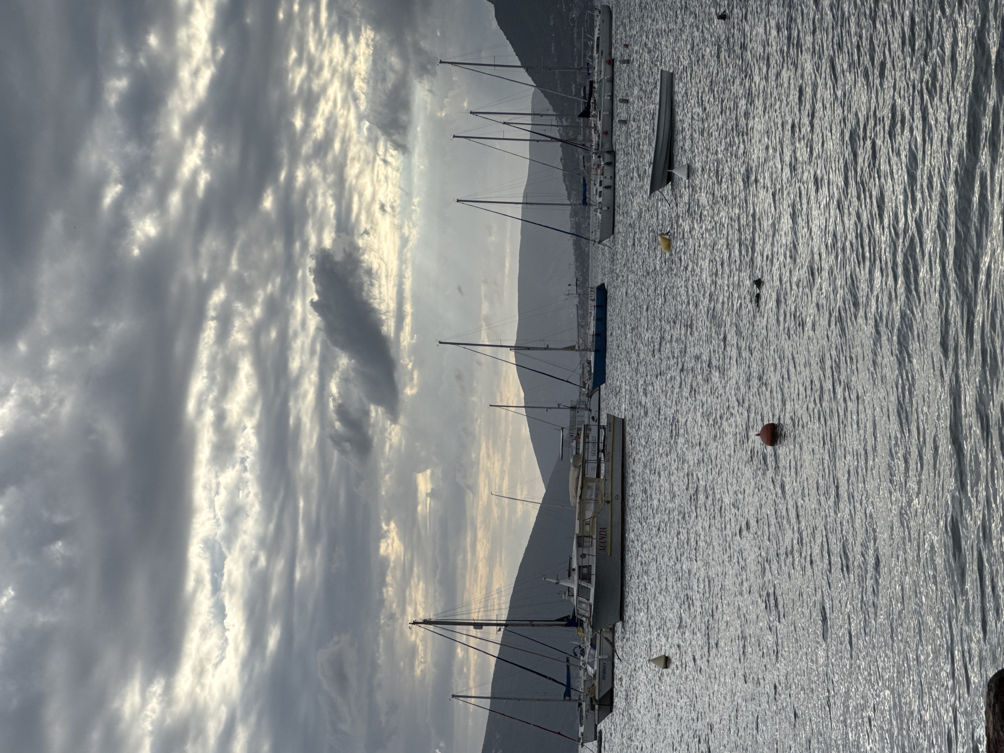 The photograph depicts a harbor scene in Tivat, Montenegro, showcasing a body of water occupied by several docked sailboats and other vessels. The background reveals a mountainous landscape partially obscured by a cloudy sky with visible sun rays breaking through. The water surface is rippled, reflecting light. Buoys are scattered across the water. There are no people visible in the frame.