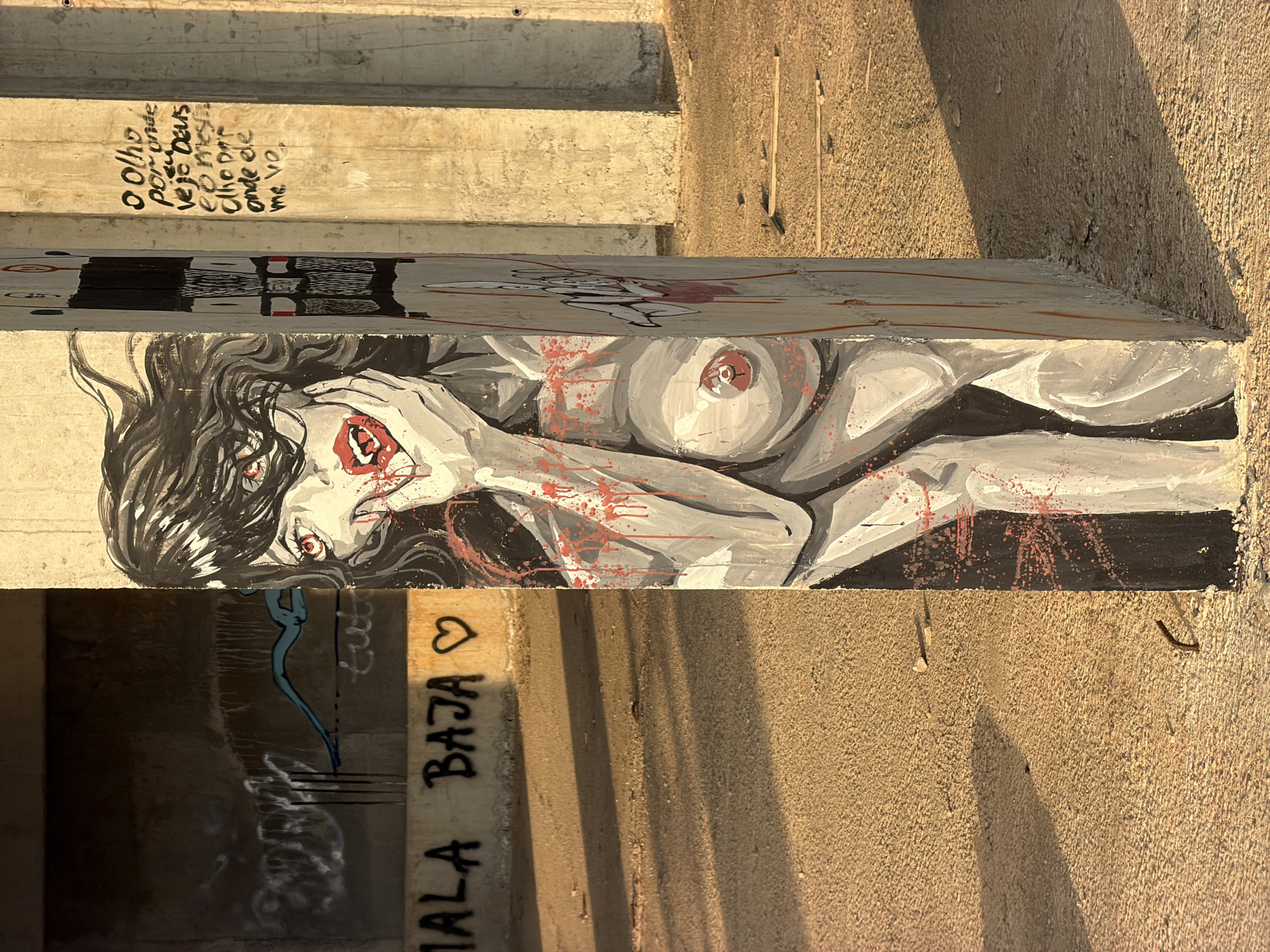 The image shows graffiti on a concrete pillar in Perazića Do, Montenegro. The graffiti depicts a stylized, nude woman with black hair, red eyes, and red lips, splattered with red paint that resembles blood. She is holding her hand to her face. The pillar itself is rough concrete. In the background, more graffiti is visible on a concrete wall and other pillars. The ground is also concrete and appears somewhat weathered, with scattered debris. The scene is illuminated by a warm, golden light, suggesting it was taken during sunrise or sunset. There is no indication of any people being present in the image.