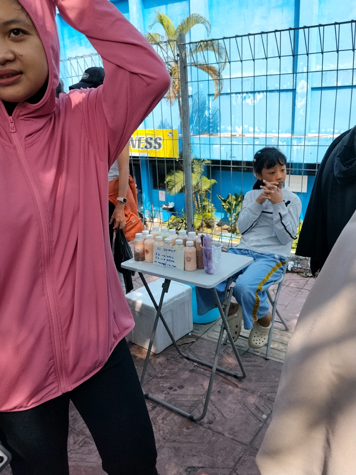 In Depok, Indonesia, a young girl sits at a small table laden with beverages, her hands clasped in front of her. She wears a light blue long-sleeved shirt and blue jeans with a yellow stripe. Behind her, a bright blue wall with palm tree murals is visible through a metal fence. To her left, a person in a pink hooded jacket is partially visible, their arm raised. The table holds several bottles of drinks and a plastic cup with a straw. The surrounding area appears to be outdoors, with a tiled ground and some foliage.