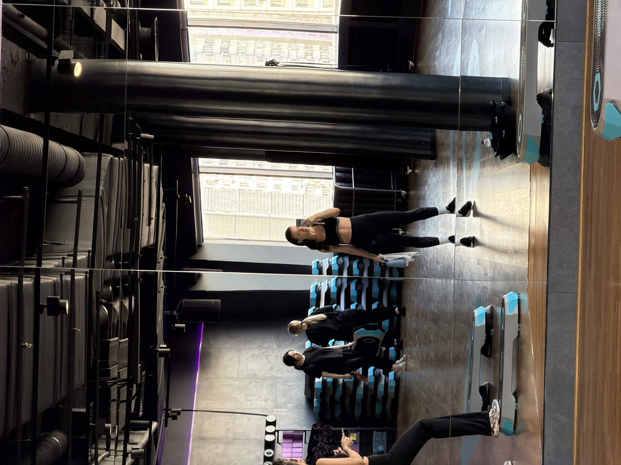 This image depicts a fitness studio located in Moscow, Russia. The scene is captured through a large, floor-to-ceiling mirror that reflects the interior and exterior environment.
**Environment:**
The studio features an industrial aesthetic with an exposed black ceiling showcasing ventilation ducts, track lighting fixtures, and large black speaker units. Walls are painted a muted concrete grey, with a horizontal purple LED light strip running along one section. Large windows on the right side of the room offer a view of multi-story residential buildings, characteristic of an urban landscape. A dark, cylindrical support pillar is visible near the windows, also reflected in the mirror. The flooring is composed of light-colored wooden panels in the foreground and a darker, possibly tiled or concrete section closer to the mirrored wall. Numerous grey and blue step platforms are stacked against a wall in the background and placed on the floor. Black rolling carts or equipment racks are also visible.
**People and Actions:**
Four individuals, all adult females, are present in the scene, dressed in athletic wear.
1. **Primary Subject (Photographer):** A woman, partially visible on the left, is taking a mirror selfie with her phone held in her left hand. She wears a black sports bra and black leggings, exposing her midriff. Her reflection, visible on the right side of the mirror, shows her holding a blue water bottle in her right hand and smiling subtly. She has dark hair, styled down.
2. **Background Figure 1:** Another woman is observed in the mid-background, walking towards the right. She wears a black t-shirt and dark pants, holding a dark bottle in her right hand. She has dark hair.
3. **Background Figure 2:** A third woman, positioned behind and to the right of Background Figure 1, is walking away from the camera's perspective. She wears a black t-shirt and dark pants, carrying a grey and blue step platform with both hands. Her hair is light-colored and pulled back into a ponytail.
4. **Background Figure 3:** A fourth woman is partially visible further to the left in the background, near the stacked step platforms. Her attire is dark.
**Objects:**
Prominent objects include:
* Several grey and blue aerobic step platforms, both stacked and individually placed on the floor.
* A blue water bottle held by the primary subject's reflection.
* A dark bottle held by Background Figure 1.
* A smartphone held by the primary subject.
* Black speaker units and track lighting fixtures on the ceiling.
* Black cylindrical pillar.
* Black rolling equipment carts.
The overall scene suggests a active fitness environment with individuals engaged in personal routines or preparing for a group activity.