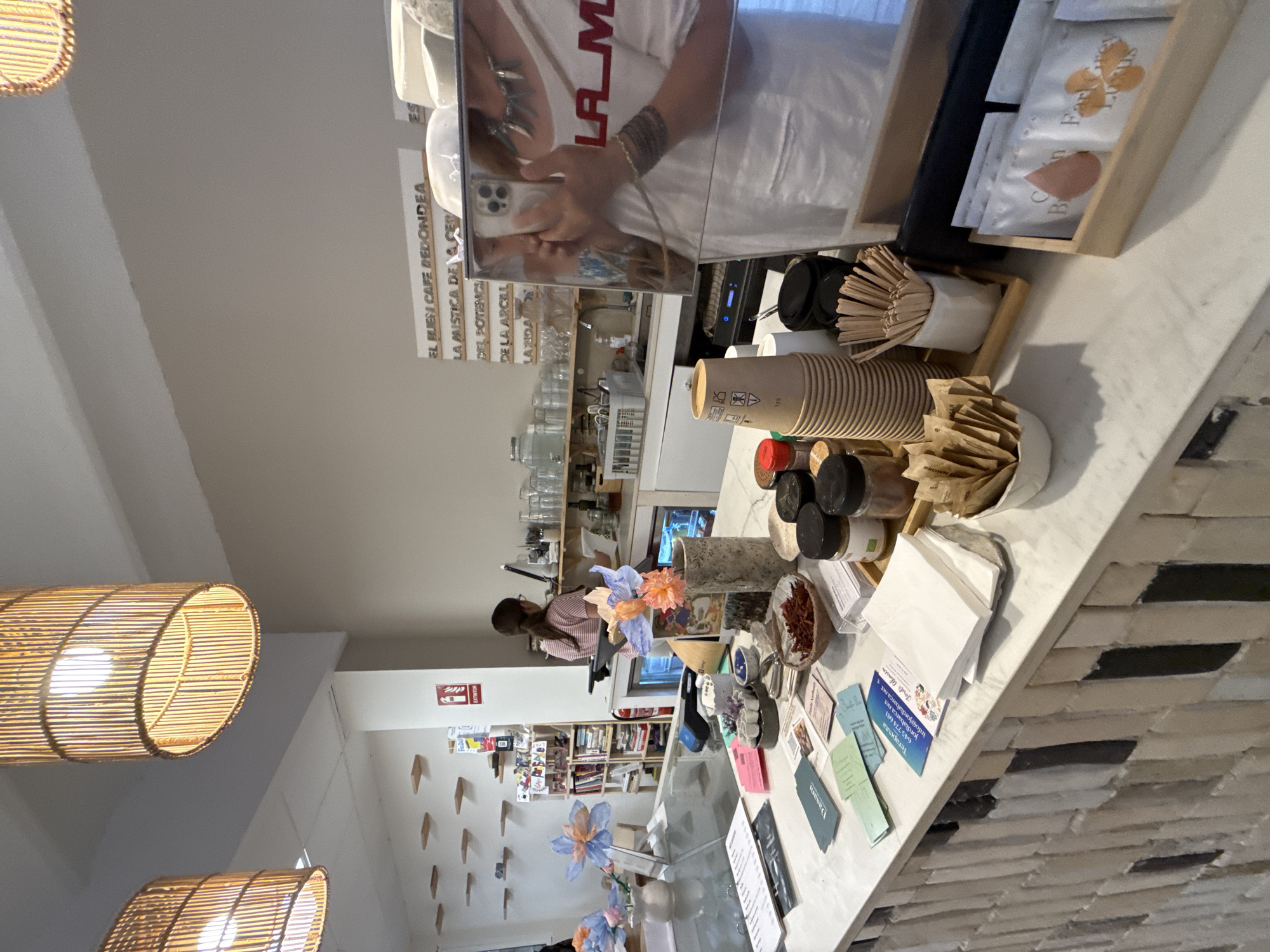 The image depicts the interior of a cafe in Mataró, Spain, with a focus on the counter area. A woman, partially visible in the reflection of a chrome espresso machine, is standing behind the counter, looking at her phone. She is wearing a white apron and a patterned shirt. The counter itself is made of white marble and is cluttered with various items, including stacks of paper cups, wooden stir sticks, small jars of spices, and packets of tea. Decorative flowers in vases are also present. Behind the counter, shelves are stocked with books, merchandise, and glassware. A red fire extinguisher is mounted on the wall. The lighting in the cafe comes from two large, cylindrical rattan lamps hanging from the ceiling. The cafe has a minimalist aesthetic with white walls and light-colored shelves. The overall impression is that of a well-lit and organized space.