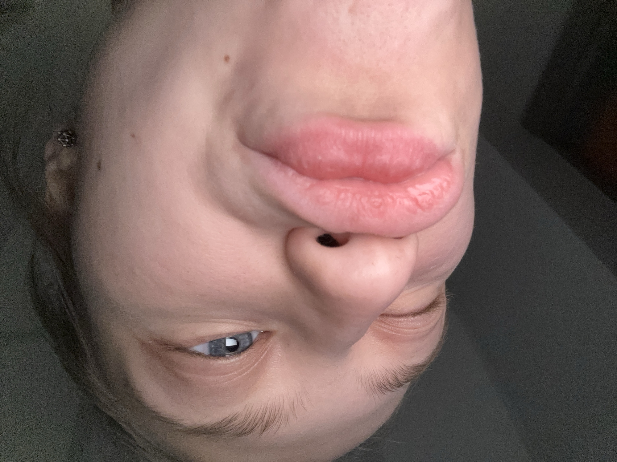 A close-up, upside-down image focuses on a person's face. The individual has fair skin, blue eyes, and light-colored hair. The mouth is prominent, and the nose is positioned directly below it. One eye is open, while the other is partially closed. An earring is visible in the ear. The background is a dark, solid color. The location in the city of Perm, Russia cannot be determined from this image.