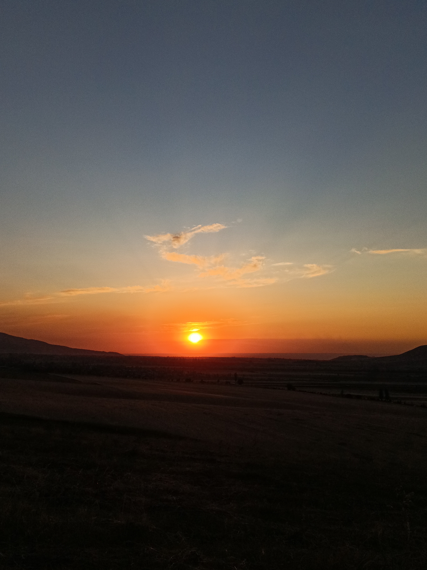 This is a photograph of a sunset over a landscape. The sun is a bright orange orb positioned in the center of the frame, casting a warm glow across the sky. The sky transitions from a deep blue at the top to shades of orange and yellow near the horizon. There are a few wispy clouds scattered across the sky. In the foreground, there is a dark, silhouetted landscape with rolling hills. The horizon line is low in the frame, with mountains visible in the distance. The scene appears to be a rural or natural setting. No people or specific objects are clearly discernible in the image due to the lighting and composition. The location is described as Arashan, Kyrgyzstan.