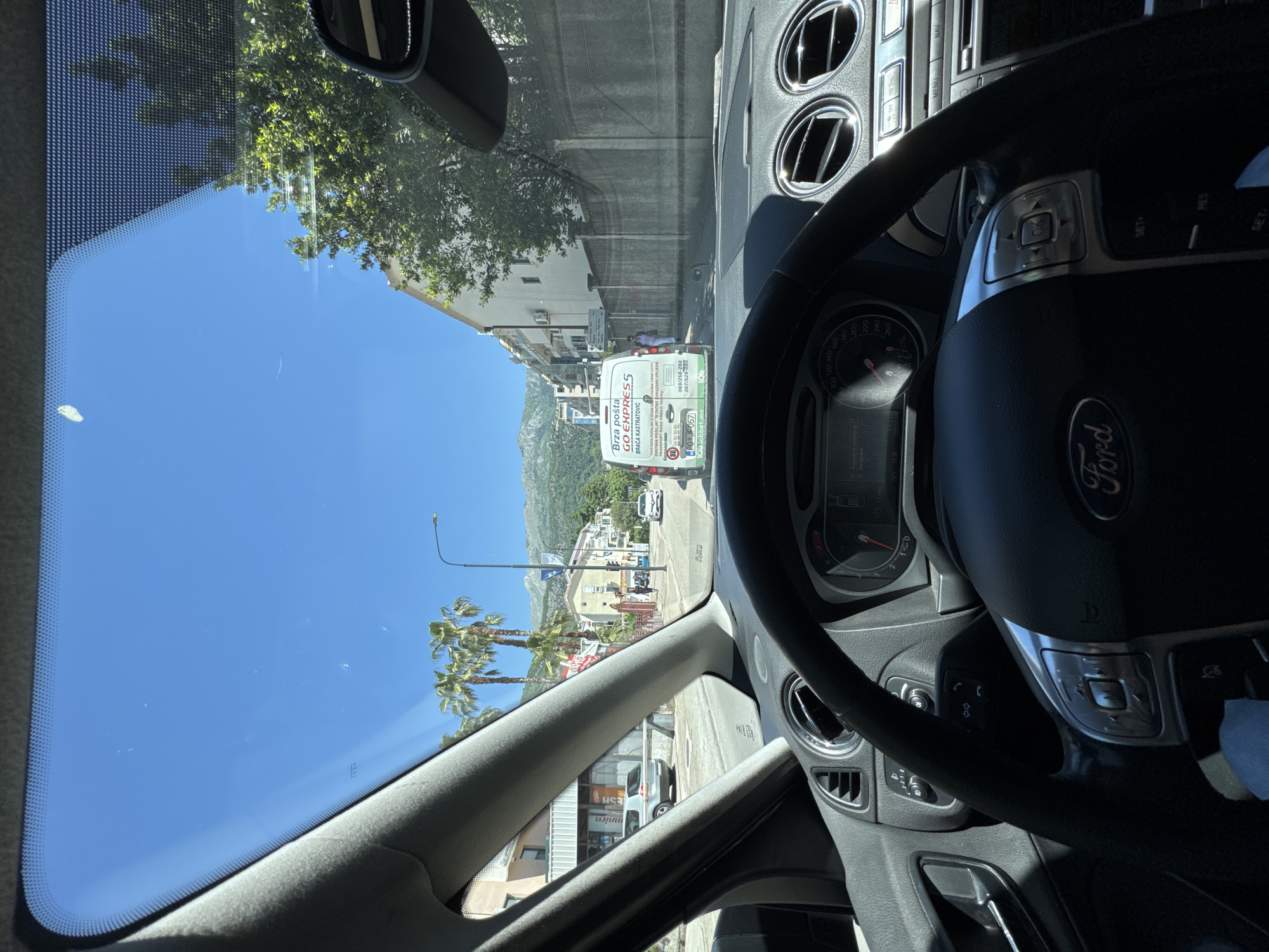 The image, taken from inside a Ford vehicle, captures a street scene in Budva, Montenegro. Through the windshield, palm trees line a street flanked by buildings, with mountains visible in the background. A white delivery van branded with "GO EXPRESS" is in the immediate vicinity. The vehicle's steering wheel and dashboard are in view, partially obstructing the street scene.