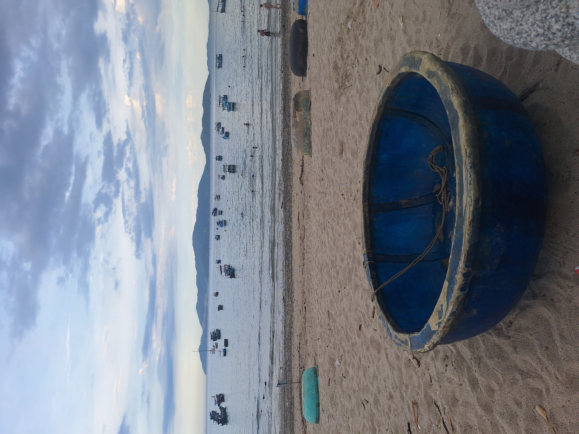 This is a photograph of a beach in Nha Trang, Vietnam. In the foreground, a large, round, blue basket boat sits on the sandy beach. It is weathered, with peeling paint and a rope inside. Further back on the beach are two other basket boats, one turquoise and one dark gray, and what appears to be the top of another blue boat peeking into the frame from the right. The beach itself is sandy with some debris scattered around.
The water is calm and stretches out to the horizon. In the distance, there are many small boats, some with canopies, floating on the sea. The background features a range of hills or mountains under a cloudy sky with patches of blue. Several people are visible in the water, wading and swimming. The overall scene depicts a typical coastal landscape with traditional fishing vessels, suggesting a working fishing village or a popular tourist beach.