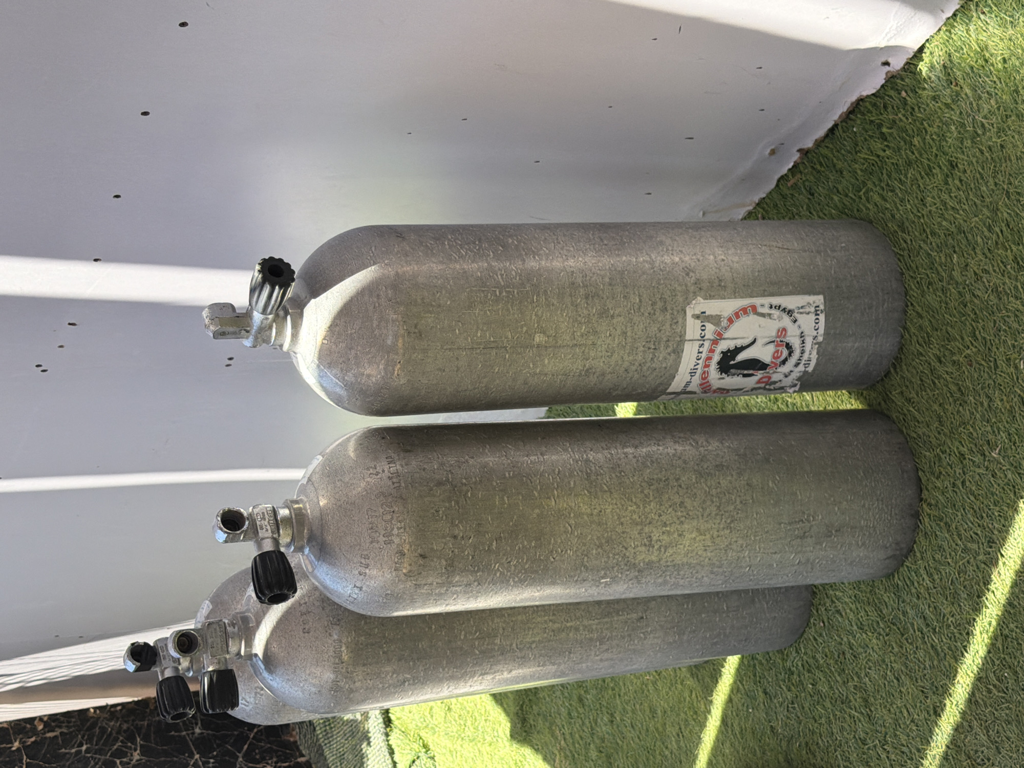 Four aluminum scuba tanks stand upright on a green, artificial turf surface, positioned in front of a textured white wall. The tanks are cylindrical and appear to be of similar size, with silver metallic finishes. Each tank is topped with a black valve mechanism. Some of the tanks have visible markings and labels, including one with a sticker that appears to be a company logo with text that reads "millennium-divers.com" and "sheikh - Egypt". The lighting suggests an outdoor setting with shadows cast across the scene, indicating daylight. The background wall has several small, dark marks. The scene is devoid of people. The location is identified as Sharm El Sheikh, Egypt.