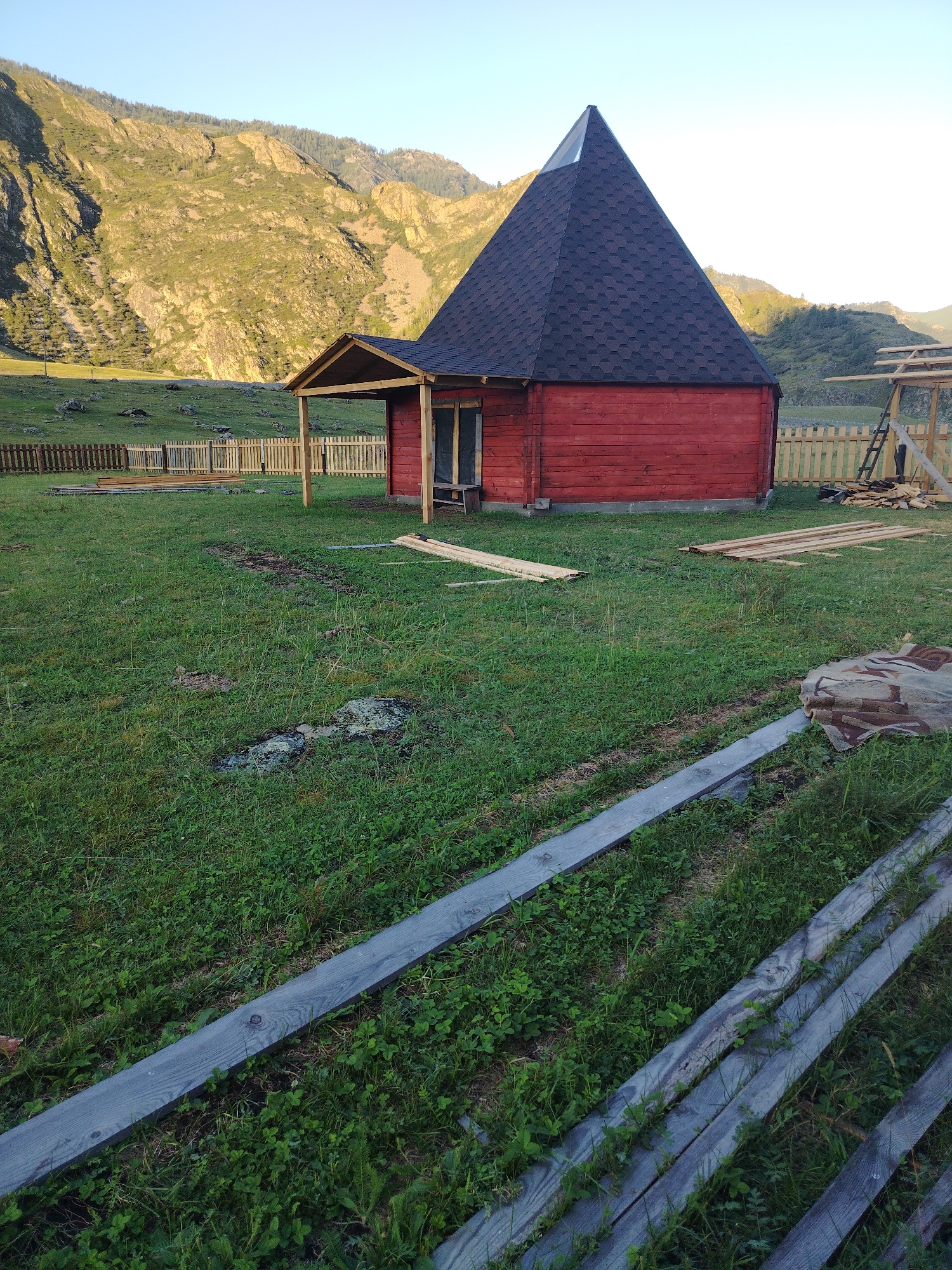 A small, red, wooden structure with a conical, dark gray roof stands in a grassy field. The building has a small covered porch with wooden posts. To the right of the structure, there is a wooden fence and some construction materials, including lumber stacked on the ground and leaning against a ladder. In the background, a large, rocky mountain with trees covers its slopes rises under a clear sky. The foreground features more wooden planks laid across the grass, and a piece of fabric or tarp is visible on the right. The scene appears to be in a rural or mountainous area, possibly a campsite or construction site.