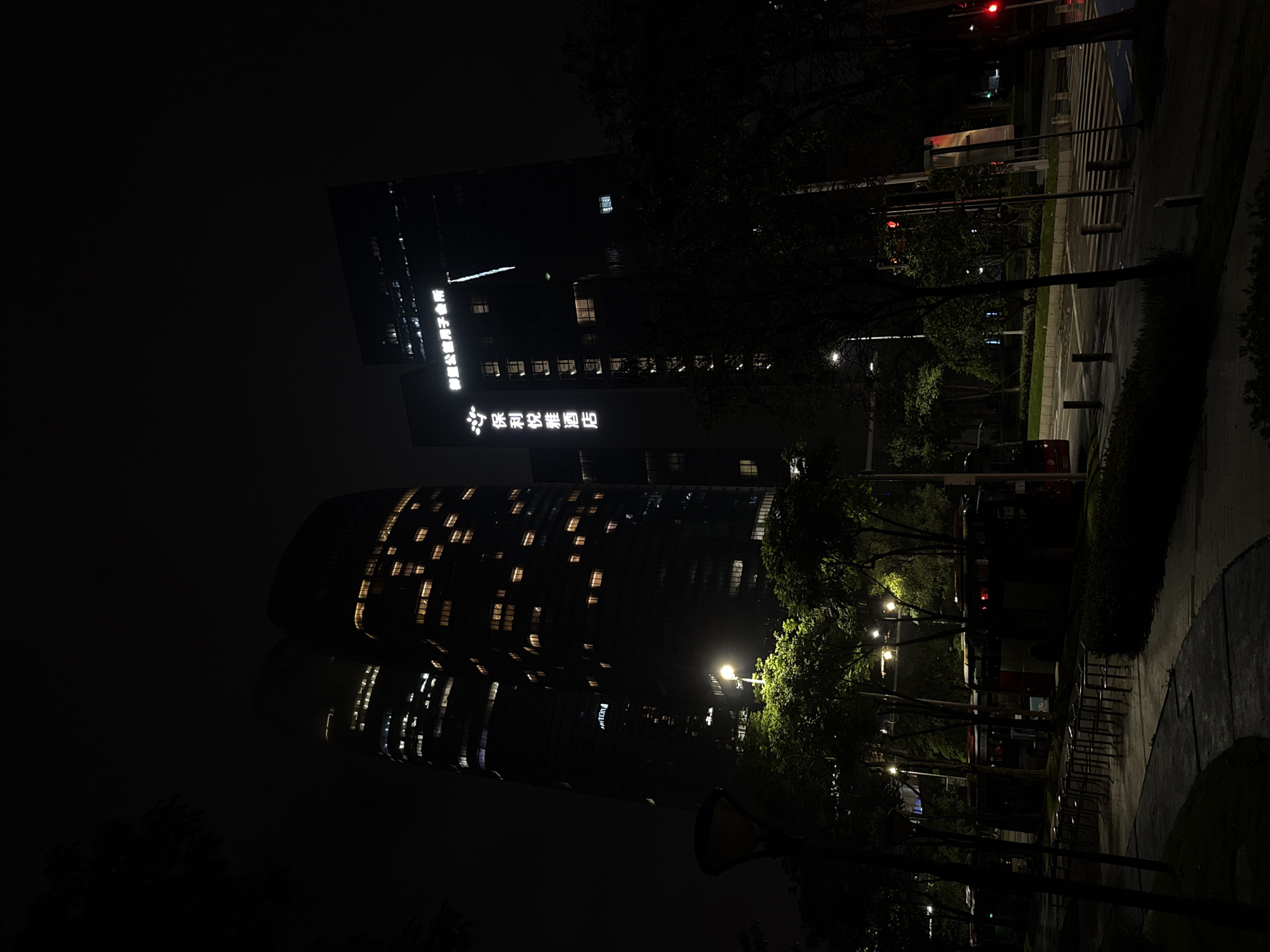 The image shows a nighttime view of two illuminated skyscrapers and a street scene in Haizhu District, China. The skyscrapers are dark glass structures with numerous lit windows, suggesting they are hotels or office buildings. One building has visible signage that reads "INTERCONTINENTAL" in English and Chinese characters, indicating it is likely the InterContinental Guangzhou Exhibition Center. The other building also has Chinese characters on its facade.
In the foreground, a street is visible with streetlights, trees, and a pedestrian walkway. Several buses are parked along the side of the street, their lights dimly visible. A crosswalk is also present on the road. The overall ambiance is urban and nocturnal, with the artificial lights of the buildings and streetlights contrasting with the dark sky. No people are clearly discernible in the image.