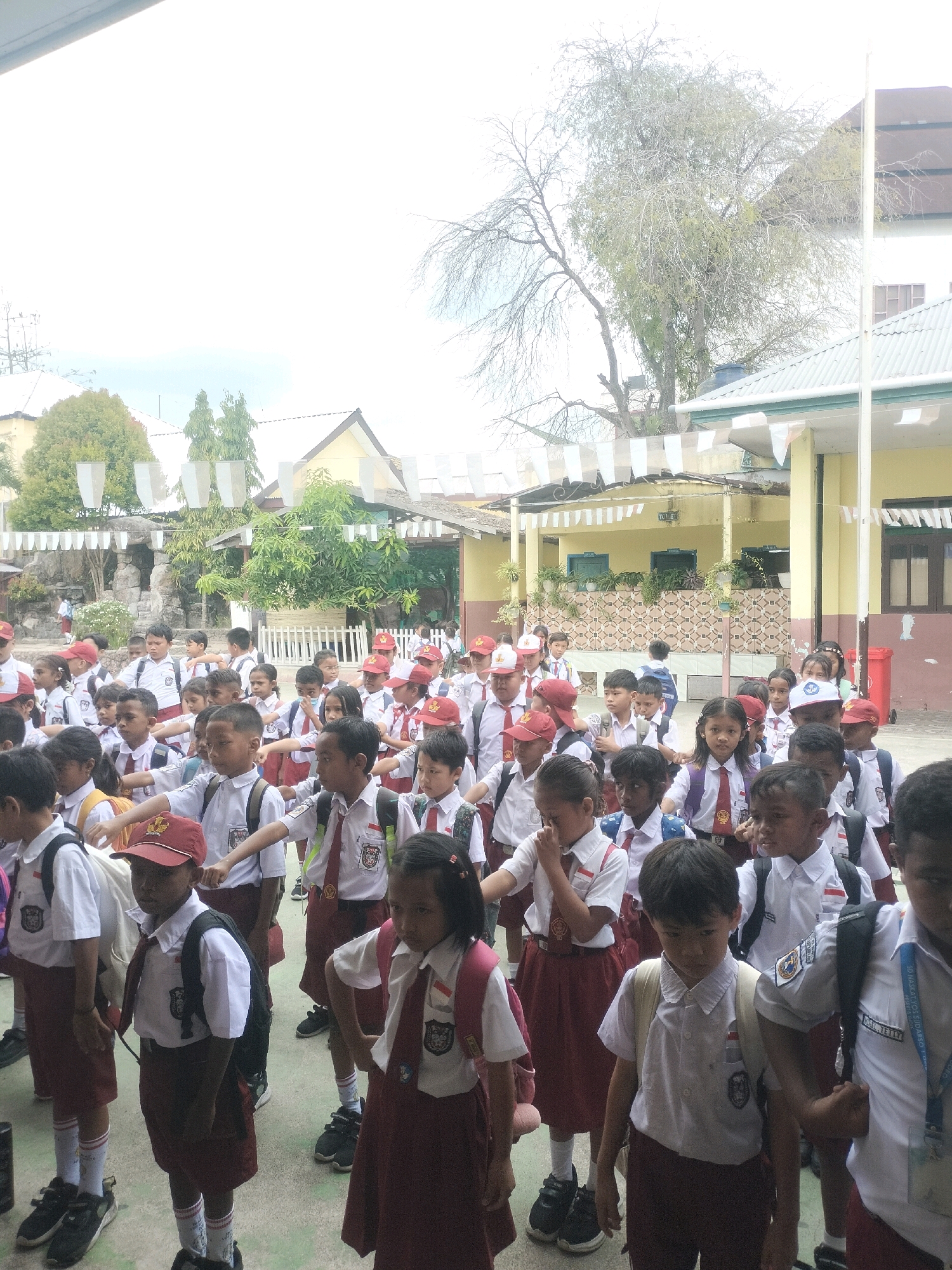 A group of Indonesian schoolchildren in uniform stand in a courtyard. The children, who appear to be of primary school age, are wearing white shirts, red ties, and red skirts or shorts. Many are also wearing backpacks. The setting appears to be a school in Dobo, Indonesia, with buildings and trees visible in the background. The children are standing in rows, and some appear to be performing a coordinated action, possibly a school assembly or a drill. The ground is paved, and there are flags or banners hanging from some of the buildings. The lighting suggests it is daytime, with bright natural light.