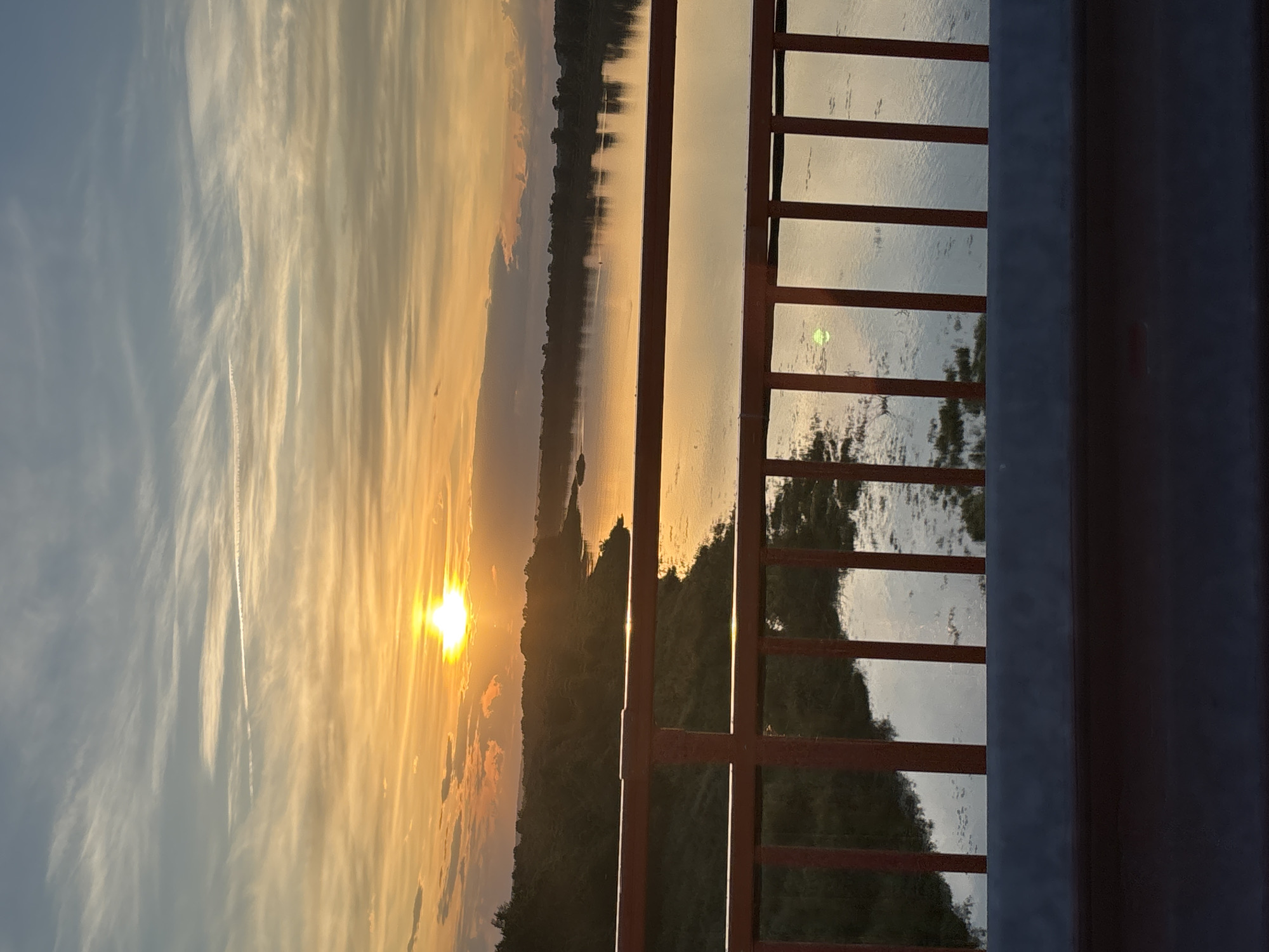 This media file captures a sunset over a body of water, possibly a river, in Diurtiuli, Russia. The scene is viewed from behind a red railing, which creates a sense of being on a balcony or bridge. The sun, a bright orb of golden light, is low on the horizon, casting a warm glow across the sky and reflecting on the water's surface. Wispy clouds streak across the sky, painted in shades of orange, yellow, and blue, indicating fair weather and a picturesque sunset.
The landscape on the opposite bank of the water is silhouetted against the vibrant sky, with the dark shapes of trees and land visible. The railing itself is composed of vertical bars and horizontal crossbars, some of which are illuminated by the setting sun. The water appears calm, with subtle ripples and reflections of the trees and sky. The overall mood is serene and tranquil, evoking a sense of peace and natural beauty. The image seems to have been taken during the golden hour, as evidenced by the warm, soft light. There is no visible text in the image.
