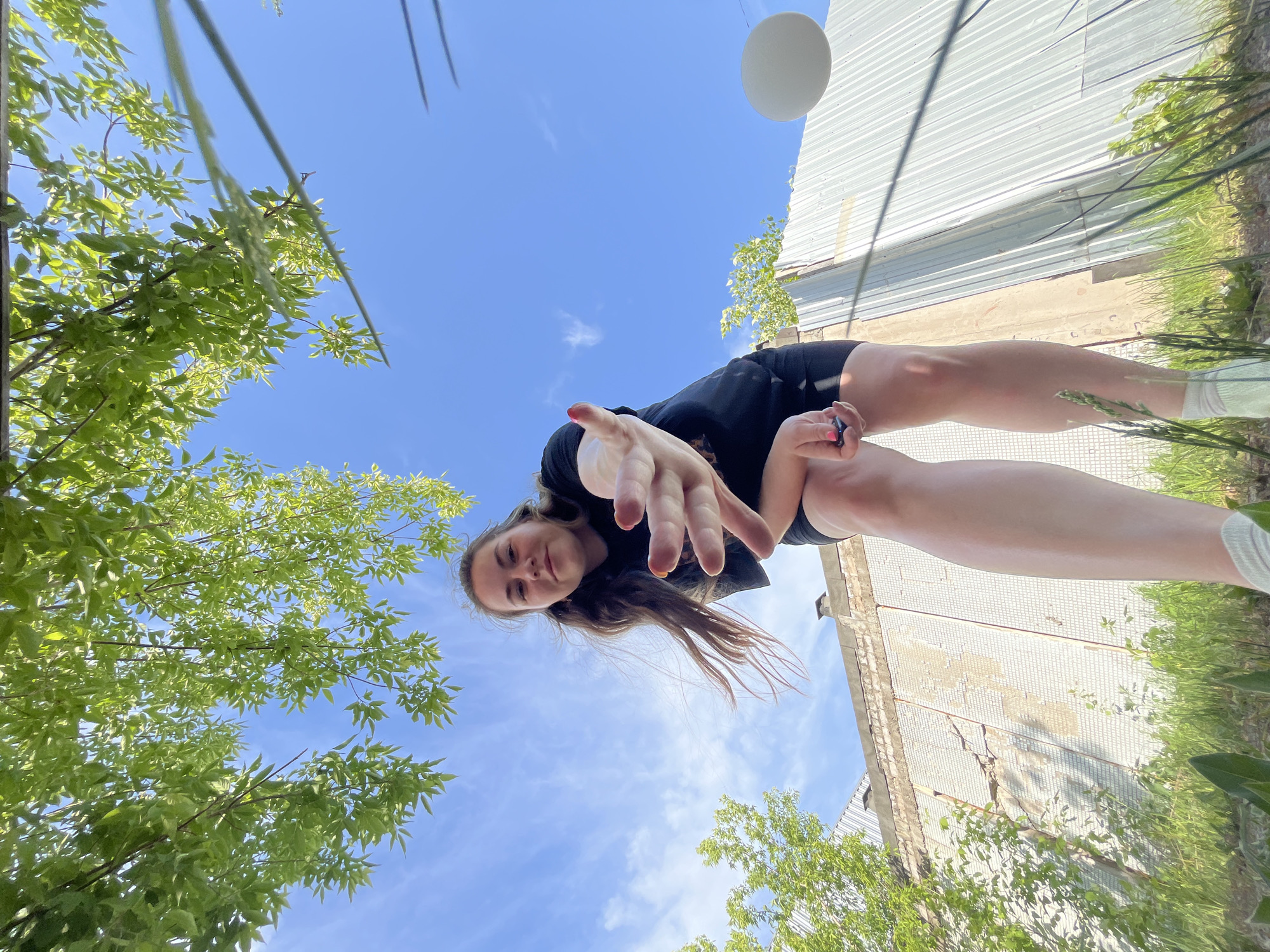 A low-angle shot shows a young woman extending her hand toward the camera. She is wearing a black shirt and shorts, and her hair is long and brown. In the background, there is a blue sky with light clouds, trees with green leaves, and a metal building. A white, round object is in the upper-right quadrant, adjacent to the metal building. Power lines can be seen at the top-left corner. The location appears to be outdoors, likely in Chaikovsky, Russia, given the request.