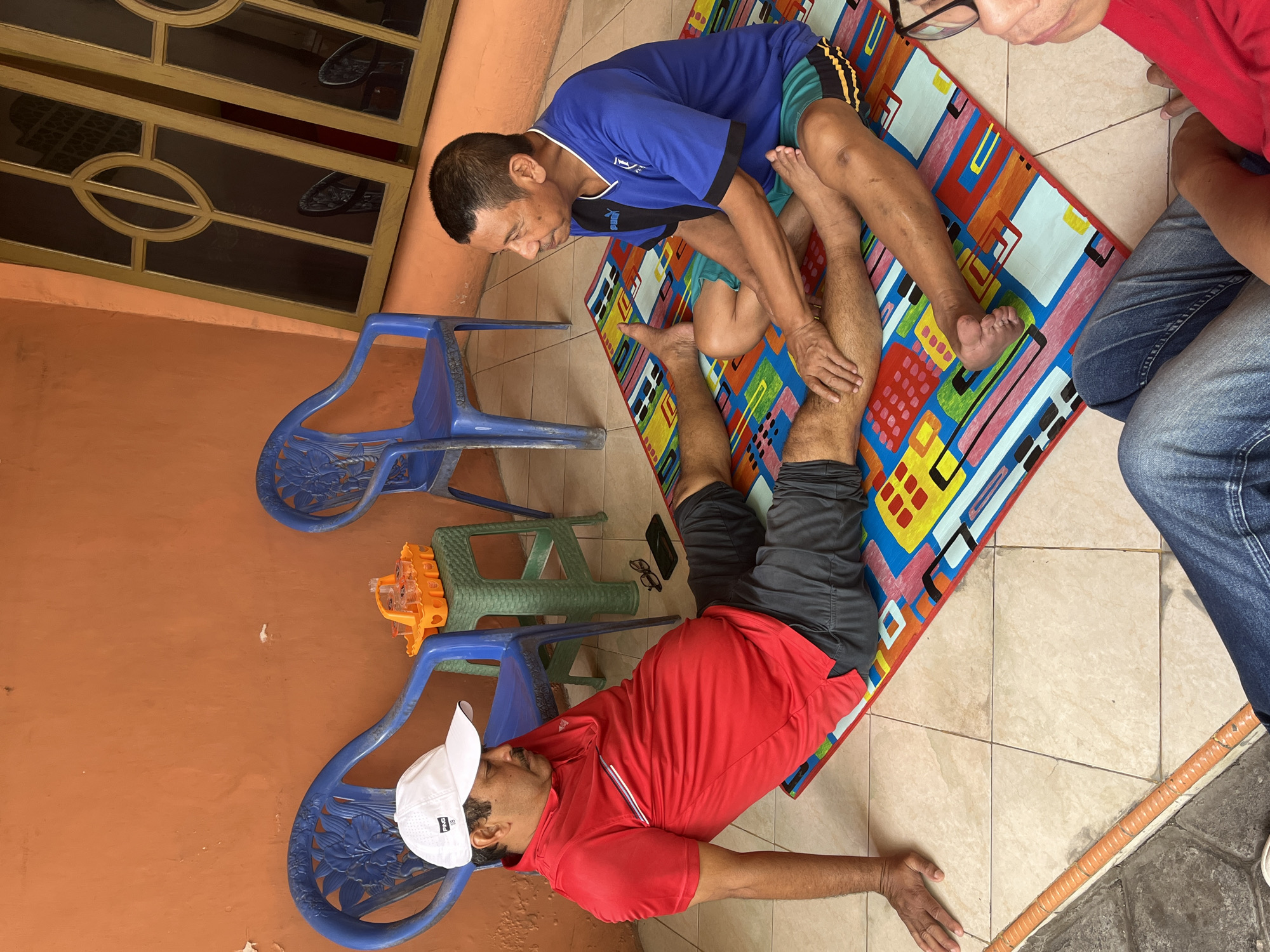 In Sidorukun, Indonesia, two men are engaged in a massage session outdoors. The man receiving the massage is seated on a colorful mat, wearing a red polo shirt and dark shorts, with a white baseball cap. He is lying on his side, with his legs extended. The man giving the massage is seated cross-legged on the mat, wearing a blue t-shirt and patterned shorts. He is actively massaging the leg of the other man.

The environment appears to be a patio or courtyard. Behind the men, there are two blue plastic chairs and an orange plastic tray holding several small cups. A portion of a building with an orange wall is visible, as is a window with a decorative wooden frame. The ground is tiled. In the foreground, another person, partially visible in blue jeans, is also seated. The overall scene suggests a casual, informal setting for a therapeutic massage.