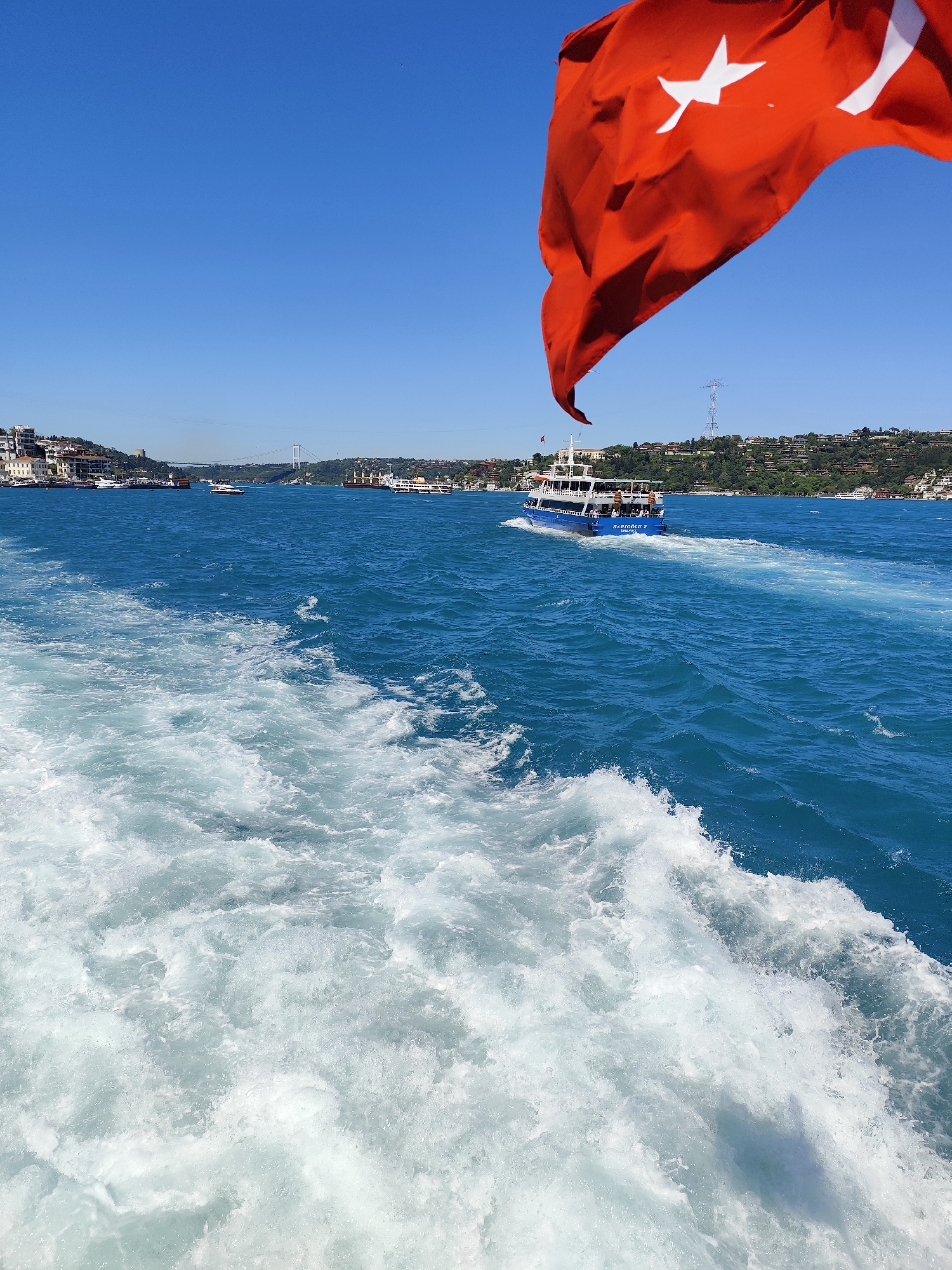 The image shows a view from a boat in the Bosphorus, likely near Beşiktaş, Turkey. The Turkish flag is prominently displayed in the foreground, partially obscuring the sky. A passenger ferry is moving through the water, creating a wake. In the background, the cityscape includes buildings and a bridge. The water is a deep blue color with foamy white waves near the camera. The sky is clear and blue. The scene is brightly lit, suggesting a sunny day.