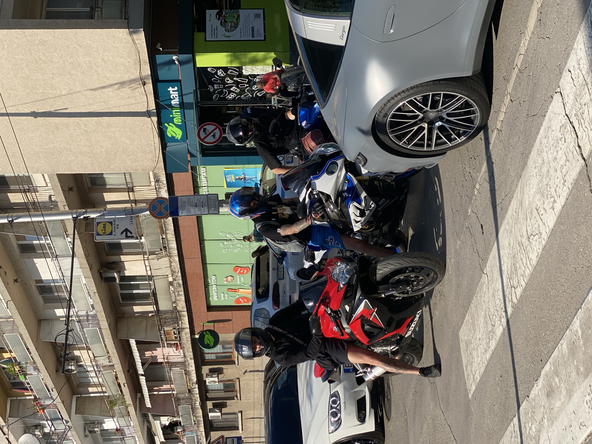 The image shows three motorcyclists parked on a pedestrian crossing in front of a Minimart store. The location appears to be a street in Pleven, Bulgaria. The motorcycles are parked between several cars. The riders are wearing helmets. Two are wearing shorts. Signage indicating Lidl is up ahead. A sign indicating no motorcycles. The weather seems sunny.