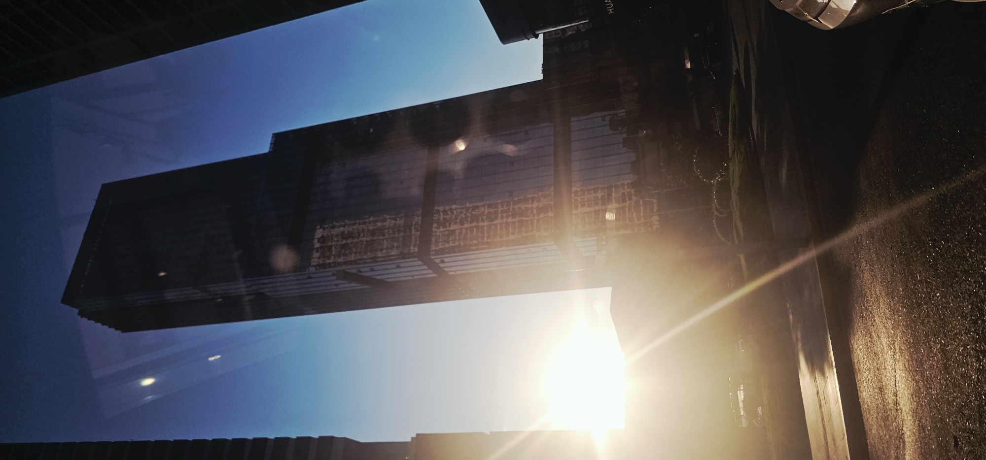 The image captures a striking perspective of a modern skyscraper in Jiulongpo District, China, during what appears to be sunset or sunrise. The tall, glass-clad building dominates the frame, its reflective surface mirroring the vibrant sky and surrounding architecture. The low angle of the sun creates a dramatic lens flare effect, casting bright rays across the scene and illuminating the asphalt road in the foreground. The street appears to be a thoroughfare, with hints of vegetation and possibly other structures in the distance. The overall impression is one of urban dynamism and the interplay of light and shadow on contemporary cityscapes.