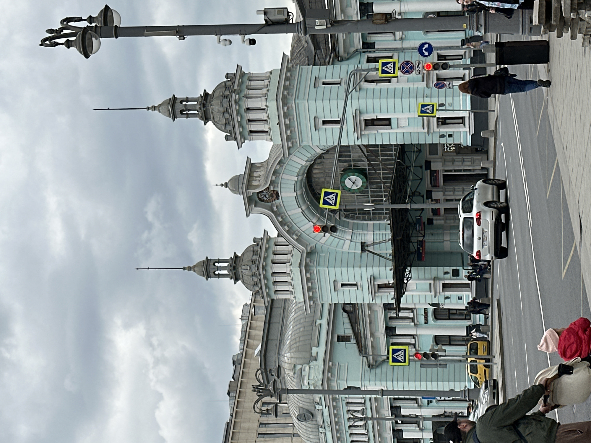 The image shows a street scene in Moscow, Russia. The Paveletsky railway station is prominently visible with its light blue and gray facade. Several cars and people are on the street. The traffic light is red. Pedestrian crossing signs are posted along the street. The sky is overcast.