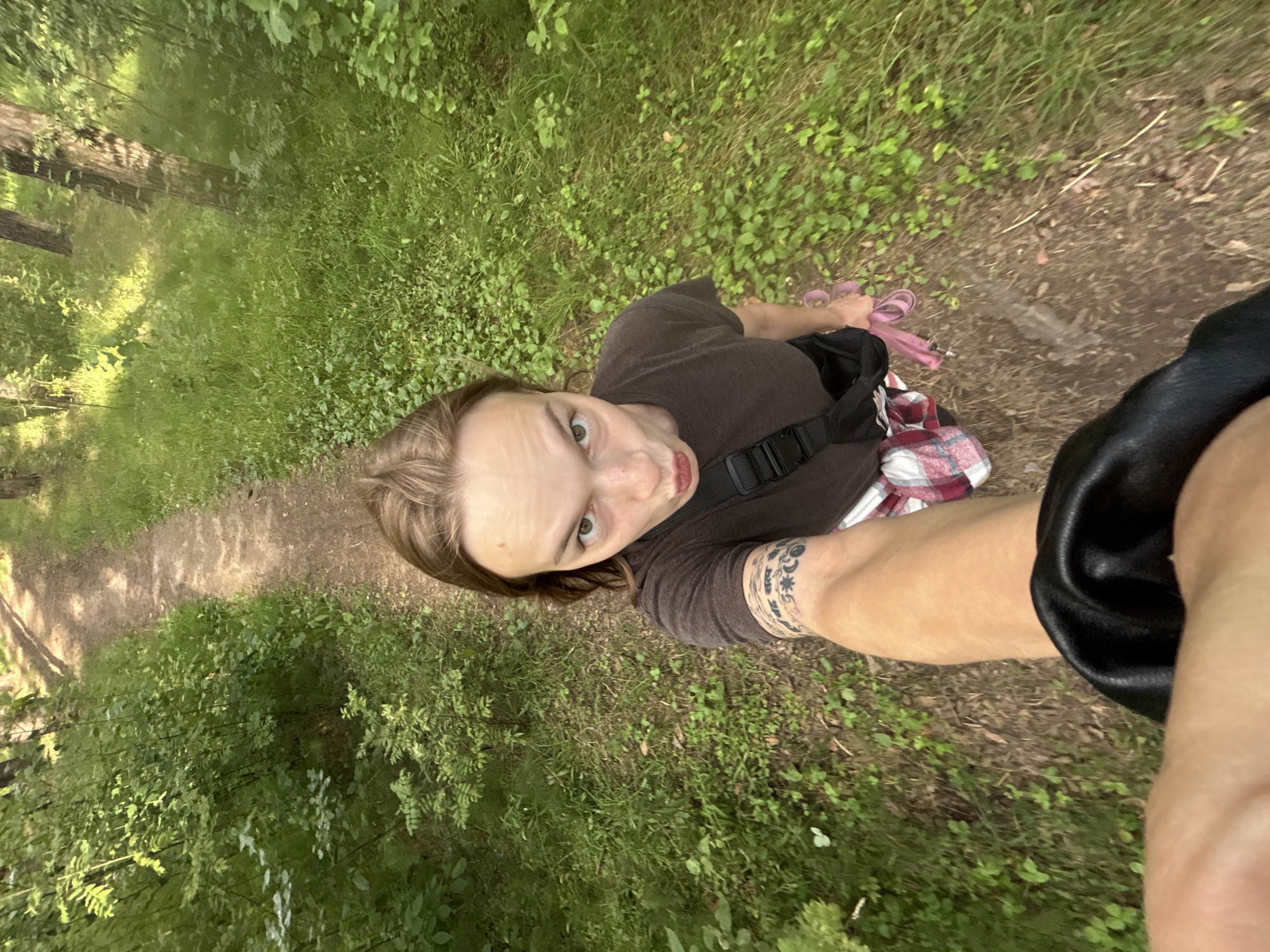 A person is taking a selfie in a forest path. The person is wearing a brown t-shirt and a plaid shirt underneath, with a black bag slung across their chest. They have tattoos on their arm and are holding a pink object, possibly a leash. The path is dirt and surrounded by lush green vegetation, with trees visible in the background. The scene is illuminated by sunlight filtering through the trees. The image was taken in Chaikovsky, Russia.