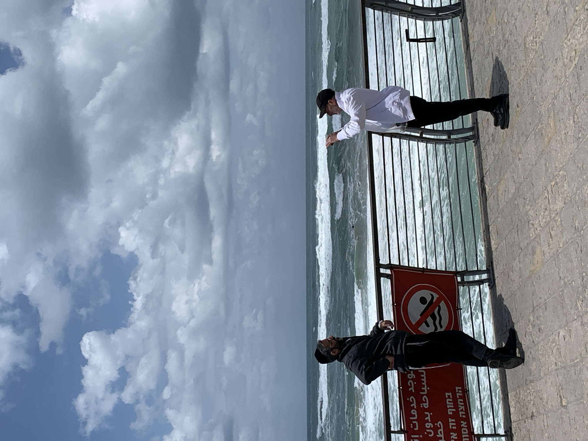 Two men are standing near a beach in Tel Aviv, Israel, on a partly cloudy day. The man on the right is wearing a white shirt, black pants, and a black baseball cap, and is looking at the water. The man on the left is wearing a black jacket, black pants, and a black hat, and is also looking at the water. A red sign indicating "no swimming" is attached to the fence behind them. The water is rough and there are waves.