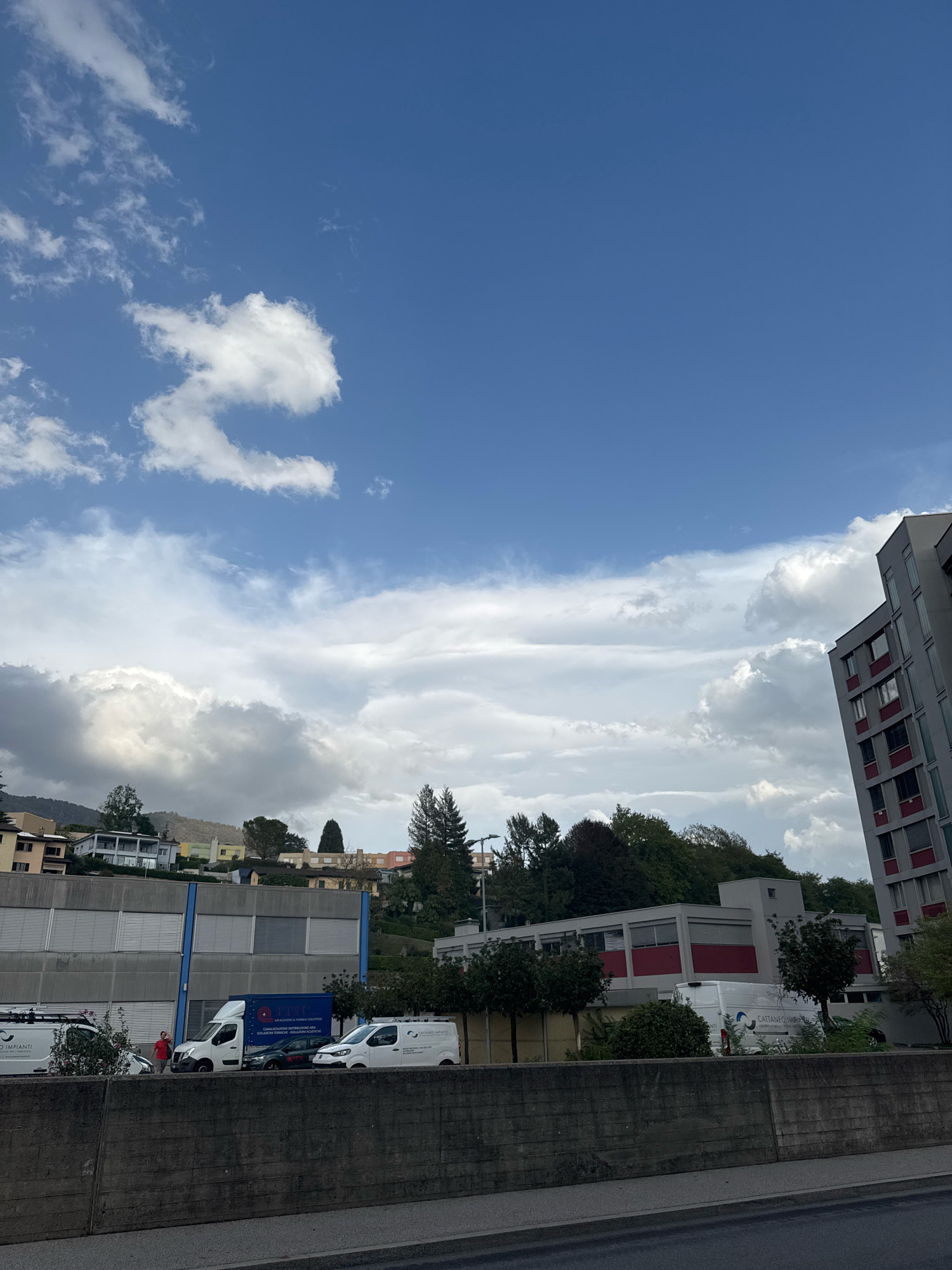 The image depicts a view of a street and buildings in Morbio Inferiore, Switzerland. In the foreground, a concrete wall runs horizontally across the frame. Behind the wall, several vans are parked, including a white van with blue lettering that reads "ITIG AIR ACOUSTIC & THERMAL SOLUTIONS". To the left of this van, a person in a red shirt is visible. Further to the left, another white van with "ATTANEO IMPIANTI" written on its side is parked.
In the midground, there are several buildings. A long, grey building with blue trim runs horizontally, and a modern building with grey facades and red accents is situated on the right side of the frame. Behind these structures, the hillside is covered with trees and houses. The sky above is a vibrant blue with scattered white clouds, some of which are dark and appear to be heavy with rain. The overall impression is of a partly cloudy day in a residential and commercial area.