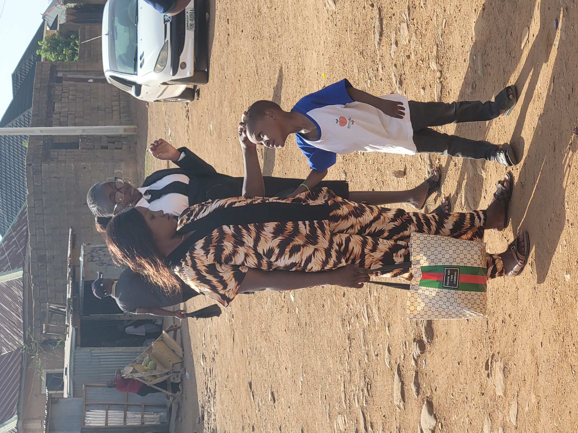 The image displays an outdoor scene on a sunny day, likely in a residential area of Jos, Nigeria, characterized by unpaved roads and simple buildings.
In the foreground, a woman with long dark hair wears a brown and black patterned dress and holds a light-colored tote bag. The bag features a repeating "GG"-like monogram pattern and a prominent green and red stripe with the visible text "THANK YOU FOR COMING". Her gaze is directed downwards. Behind her, another woman, wearing a white shirt, black tie, and black blazer, with glasses and short greyish hair, looks forward.
To the right of these women is a young boy in a white t-shirt with blue sleeves and dark trousers, wearing sandals. He is seen gently touching the head or hair of the woman in the patterned dress with his right hand, displaying a slight smile.
The ground is a dusty, sandy-brown dirt road with scattered rocks. Strong, elongated shadows cast by the subjects indicate bright daylight, suggesting mid-morning or afternoon.
In the background, several buildings constructed from concrete blocks and topped with corrugated iron roofs are visible. A white car, resembling a Peugeot 206, is parked in the upper left background, with its license plate "SHI-818JA" partially legible. Further to the left, a man in a dark t-shirt and cap is partially seen, and another person in a red shirt stands near a wooden cart that appears to hold produce. The scene captures a casual interaction among people on a typical street in the region.