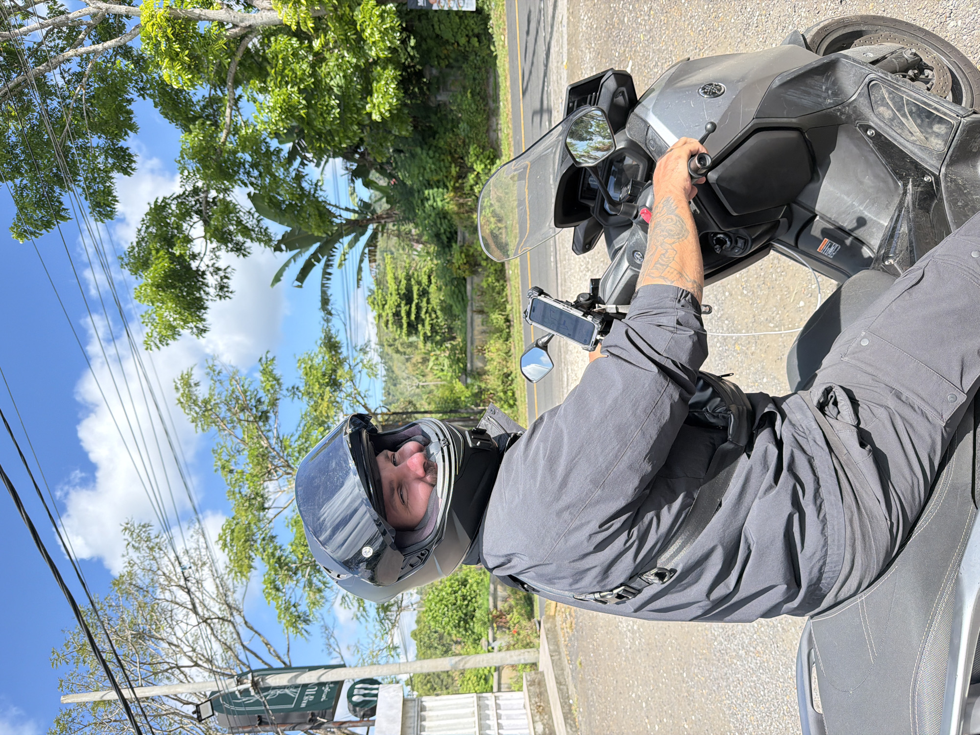 A man wearing a black helmet and a dark grey outfit sits on a grey Yamaha scooter. He is looking to his right and smiling. The scooter has a black and clear windshield, a phone mount with a phone attached, and mirrors. The man has tattoos on his left arm. The scooter is parked on a gravel road, with a paved road visible behind it. Lush green trees and vegetation are present in the background, along with a blue sky with white clouds. A pole with a sign that appears to say "HOUSE OF JU" is visible to the left of the scooter. The location is identified as Baturiti, Indonesia.