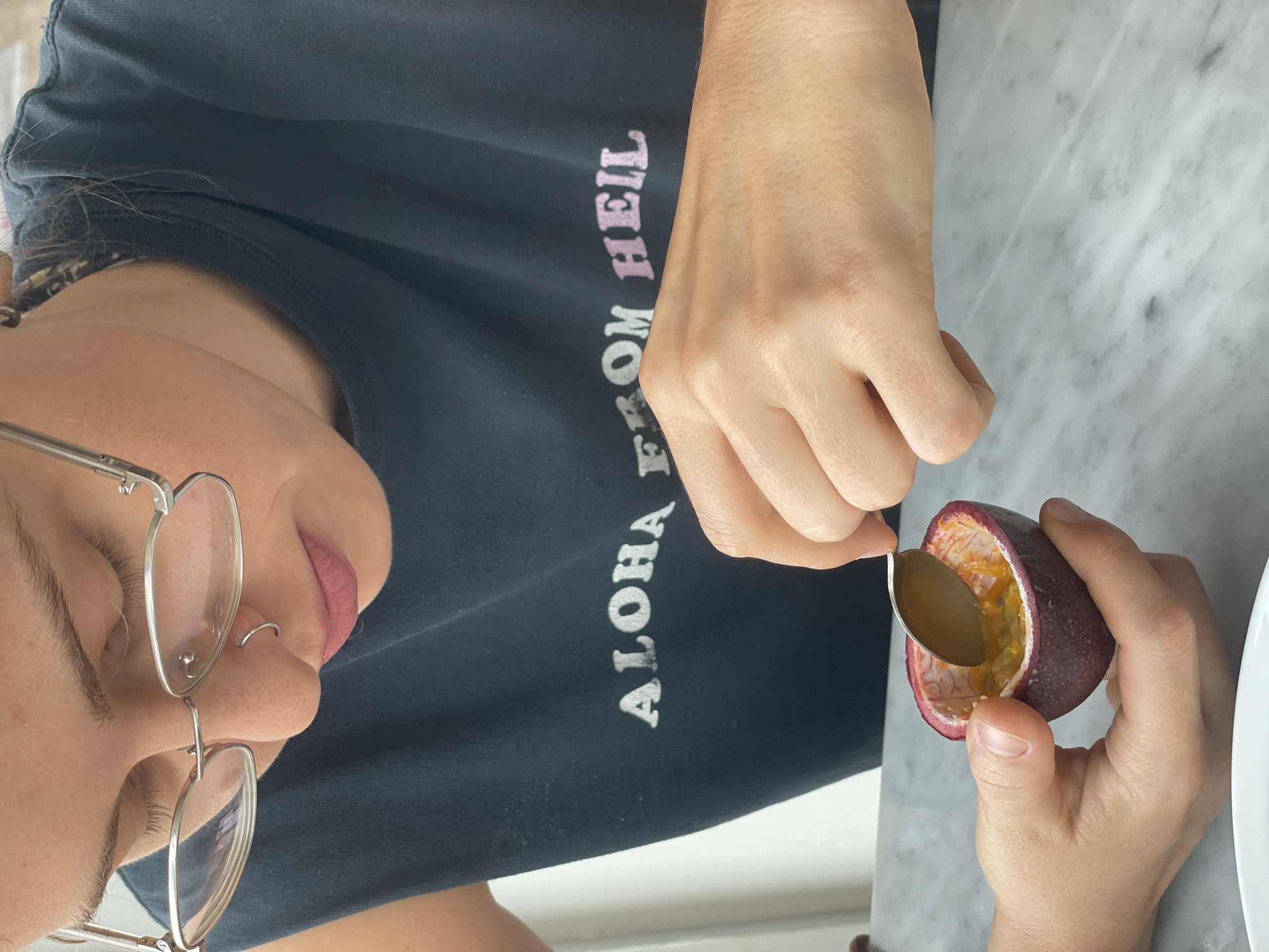 A woman with fair skin, wearing silver-rimmed glasses and a black t-shirt with the text "ALOHA FROM HELL" printed on it, is seated at a white marble table. She is holding half of a passion fruit in one hand and a small spoon in the other, apparently scooping out the fruit's pulp. The image provides no visual cues to confirm its location within Bangkok, Thailand.