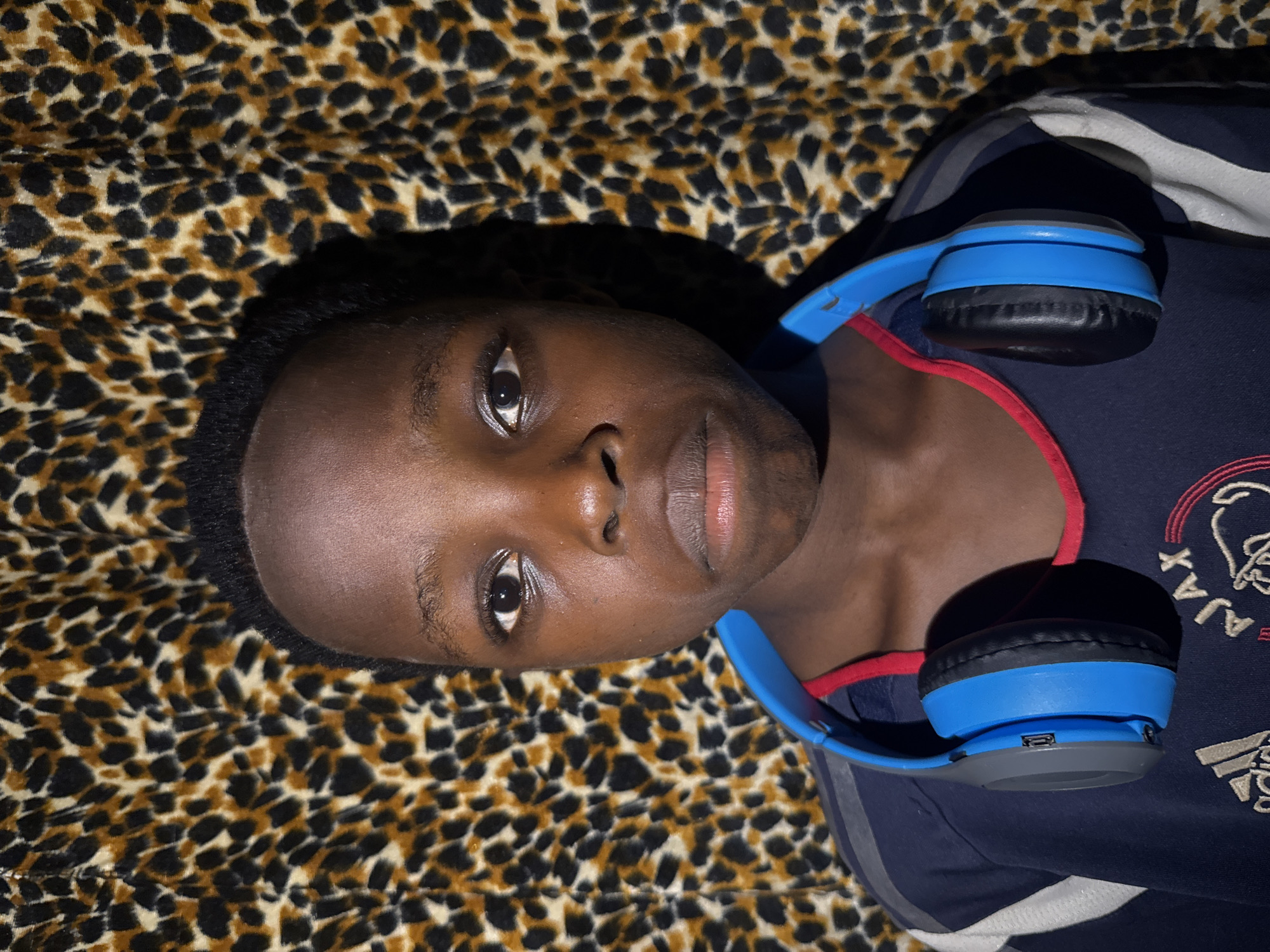 A person with dark skin and short black hair is lying on a leopard-print fabric. They are wearing a navy blue t-shirt with white stripes on the sleeves and a red collar. Blue headphones are draped around their neck. The t-shirt also features white text "AJAX" and the Adidas logo. The environment appears to be indoors, under artificial lighting. The image does not provide information to confirm location in Onitsha, Nigeria.