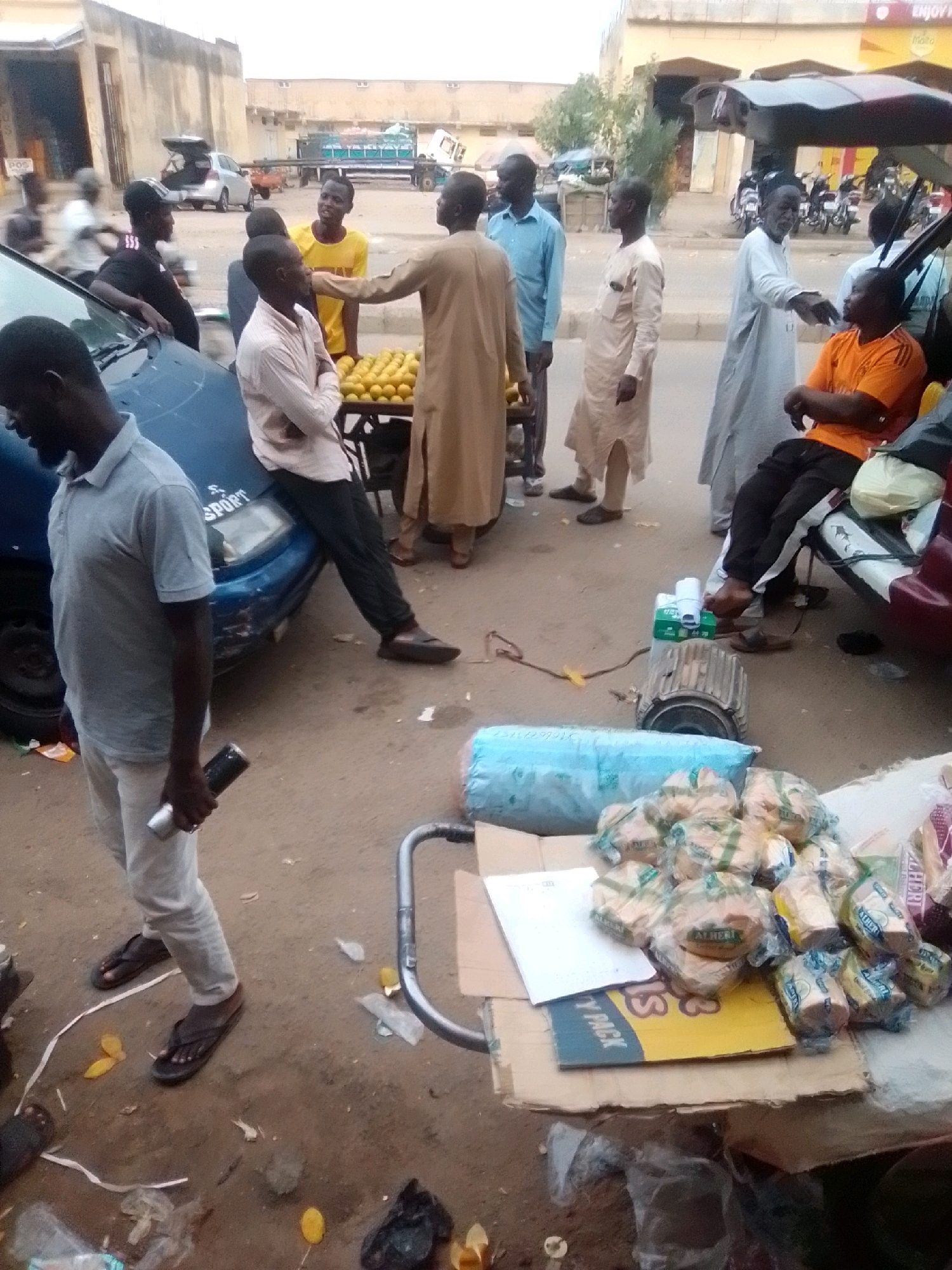 Arawa, Nigeria. A street scene depicts a group of men interacting around a parked blue car and a stall displaying oranges. One man in a light blue polo shirt and light-colored pants holds a silver cylindrical object and appears to be talking to another man dressed in a tan robe. To the right, a man in a light blue shirt and dark pants stands near a display of oranges on a table. Further to the right, an older man in a white robe gestures towards another man seated in an orange shirt. A large industrial fan or motor is visible on the ground near a box filled with packaged bread. The environment appears to be an outdoor market or roadside area with buildings and vehicles in the background.