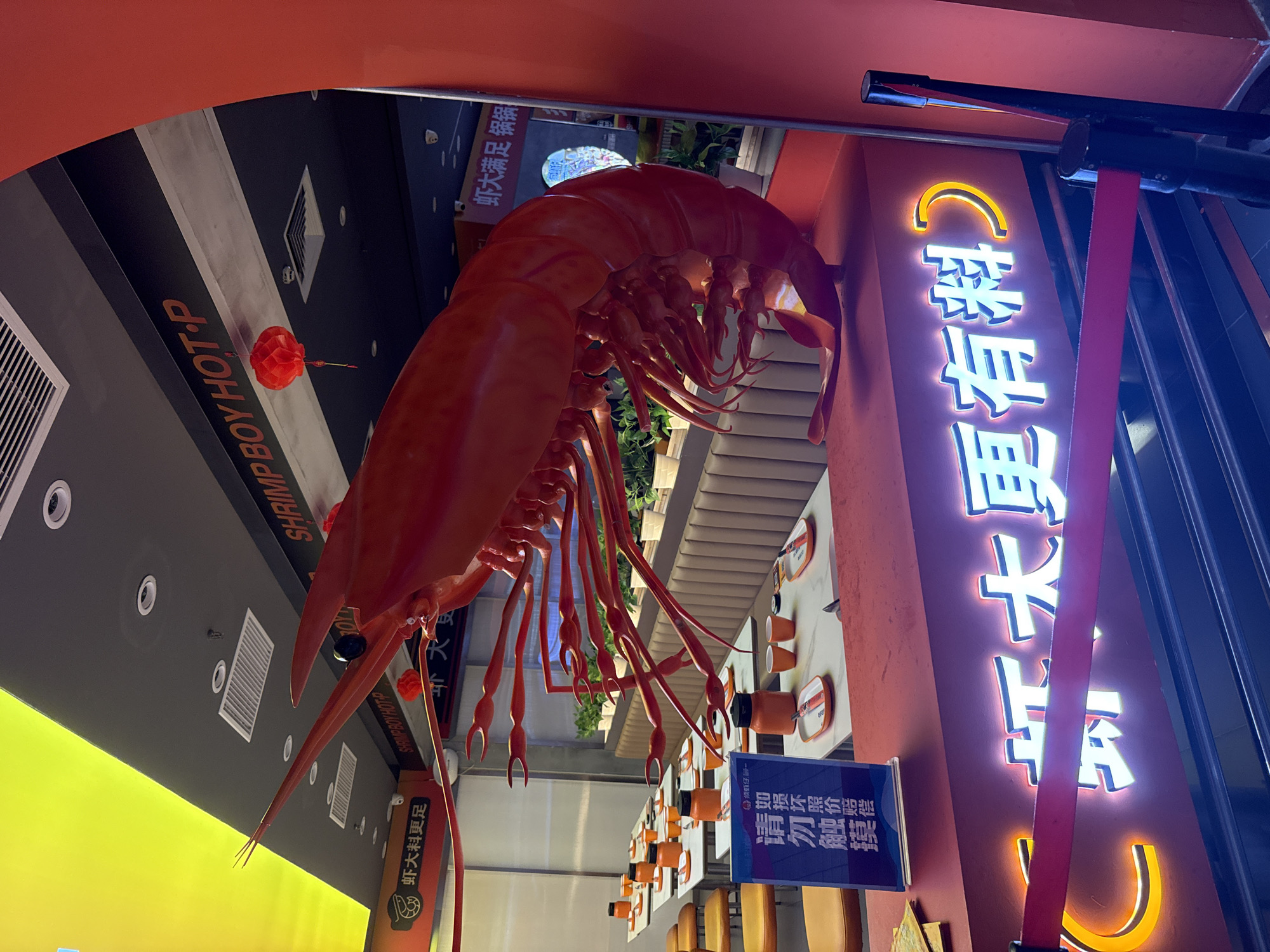The image depicts the interior of a restaurant, likely a hot pot establishment, in Huizhou, China. A large, vibrant red sculpture of a prawn dominates the foreground, positioned above a brightly lit neon sign that spells out Chinese characters. The sign's text, "虾大更有料" (Xiā dà gèng yǒu liào), suggests "Shrimp Big More Delicious" or "Shrimp Big More Ingredients."
The restaurant features several dining tables, each set with orange bowls, cups, and placemats, indicating a readiness for customers. Yellow chairs are visible, and a plush beige booth seating runs along the back wall. In the background, a sign above the tables reads "SHRIMP BOY HOT-P," confirming the establishment's theme. A small red lantern hangs from the ceiling near the "SHRIMP BOY HOT-P" sign, adding a decorative element.
A blue sign with Chinese characters is placed on one of the tables, possibly providing information or instructions. The overall ambiance is modern and inviting, with a focus on the restaurant's seafood specialty, particularly shrimp. The lighting is a mix of ambient ceiling lights and the prominent neon glow from the main sign. The restaurant appears to be empty of customers at the time the photo was taken.