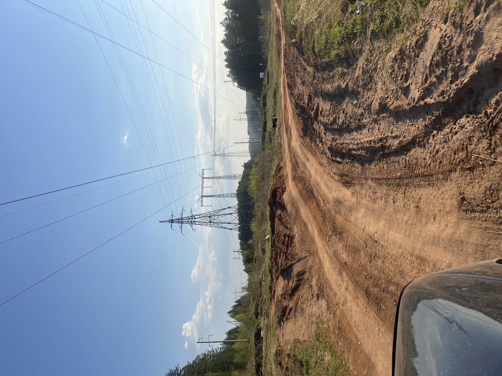 The image depicts a dirt road in a rural area. Power lines stretch across the left side of the frame, supported by metal towers. The road is lined with vegetation on the right. The top right of the image shows some tall grass. Tire tracks are visible in the dirt. Part of the vehicle the picture was taken from is visible in the bottom-right of the frame. The sky is blue with a few clouds.
The scene is located near Chaikovsky, Russia.