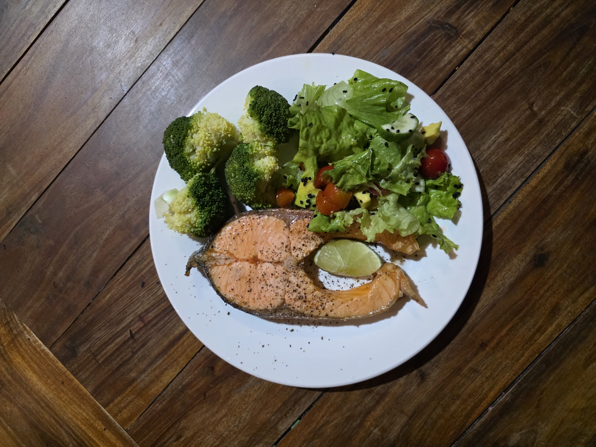 A high-angle, medium shot shows a white plate filled with food on a wooden surface. The main element of the dish is a slice of salmon, seasoned with black pepper and accompanied by a wedge of lime. Beside the salmon is a serving of broccoli and a salad with lettuce, cherry tomatoes, cucumber, avocado, and black seeds. The wooden surface appears to be a table. The food is likely a meal prepared or served at a restaurant. The location appears to be Mũi Né, Vietnam.