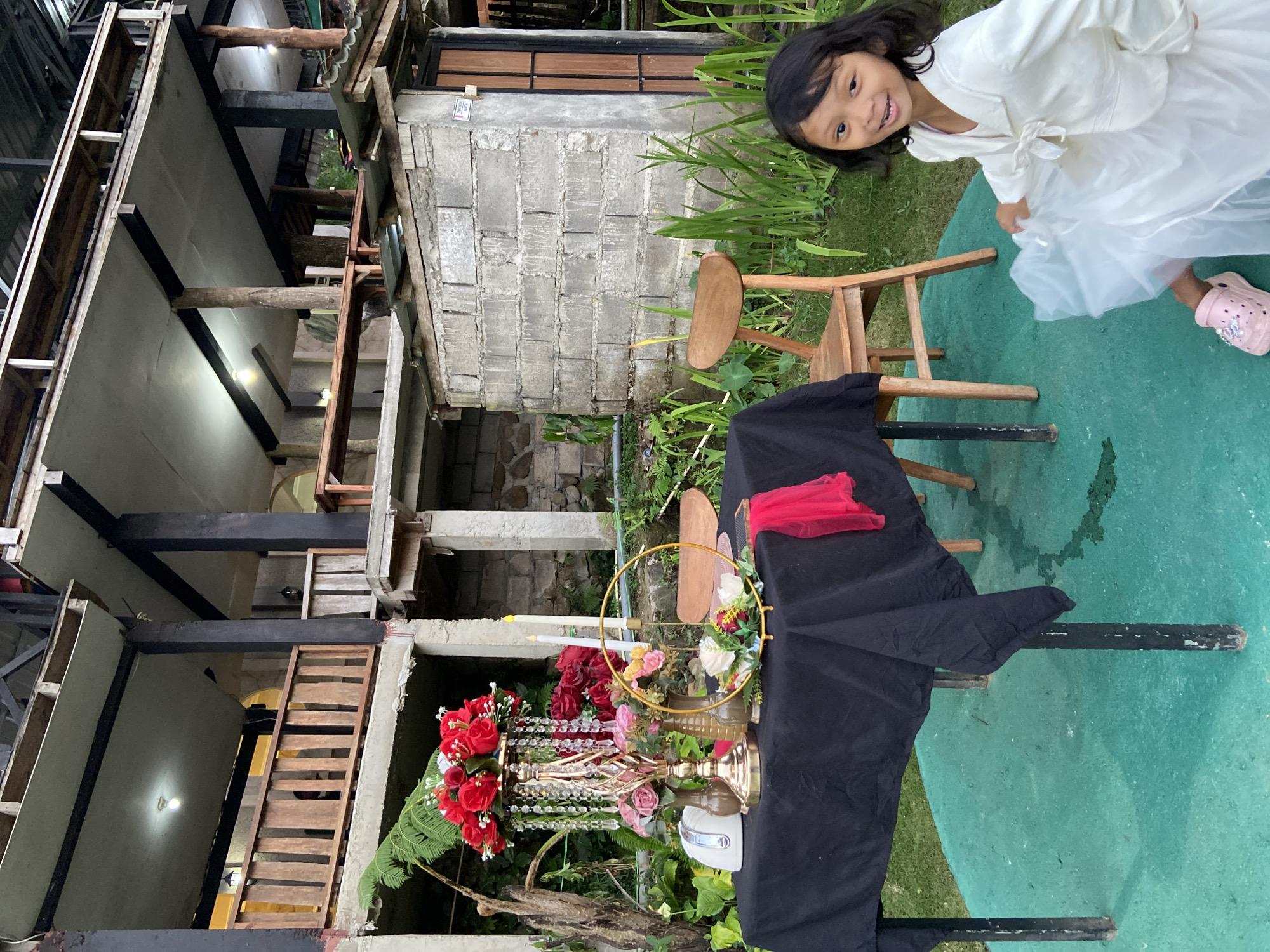In Kesiman, Indonesia, a young girl in a white dress and pink Crocs is posing playfully in front of a decorated table. The table is draped with a black cloth and adorned with a floral centerpiece featuring red and pink roses, crystal strands, and gold vases. Two white candles and a gold circular decoration with flowers are also present on the table. A wooden chair sits beside the table, and behind it, a two-story building with wooden balconies and unfinished concrete walls stands against a backdrop of lush greenery. The overall scene suggests an outdoor setting, possibly a restaurant or event space.