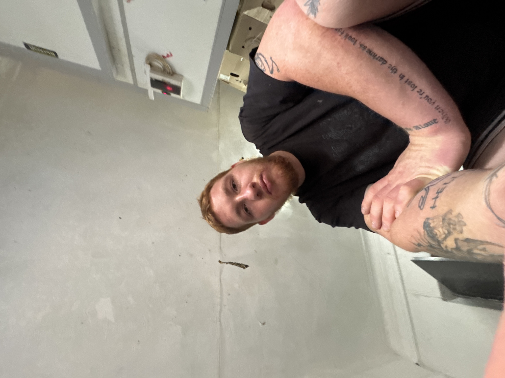 A fair-skinned male with short, red hair and a beard is shown from a low angle, looking towards the camera. He is wearing a black t-shirt. His left arm is bent, and his right arm is extended, bent at the elbow, with his forearm resting on his upper arm. The subject has multiple tattoos on his arms, including text that reads "When you're lost in the darkness, look for me." The background consists of a white, unfinished ceiling, a white wall with some fixtures, and a dark opening that appears to be a doorway. The overall lighting is dim. The location is unspecified.