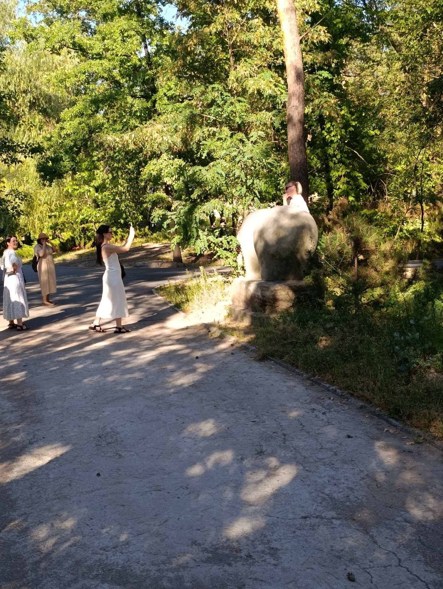In Unknown, Unknown, a paved pathway winds through a sun-dappled park setting, lined with lush green trees. Three women are visible on the pathway. The woman closest to the camera on the left wears a white patterned dress and sandals. Next to her, a woman in a light-colored dress and sandals holds her hands to her face. The third woman, in a white dress and sandals, is in the foreground, holding up a mobile phone to take a photo or video. In the background, near a large, rounded stone sculpture, a fourth person, possibly a man wearing a light-colored shirt, is partially visible. The scene is characterized by natural light filtering through the trees, creating shadows on the ground.