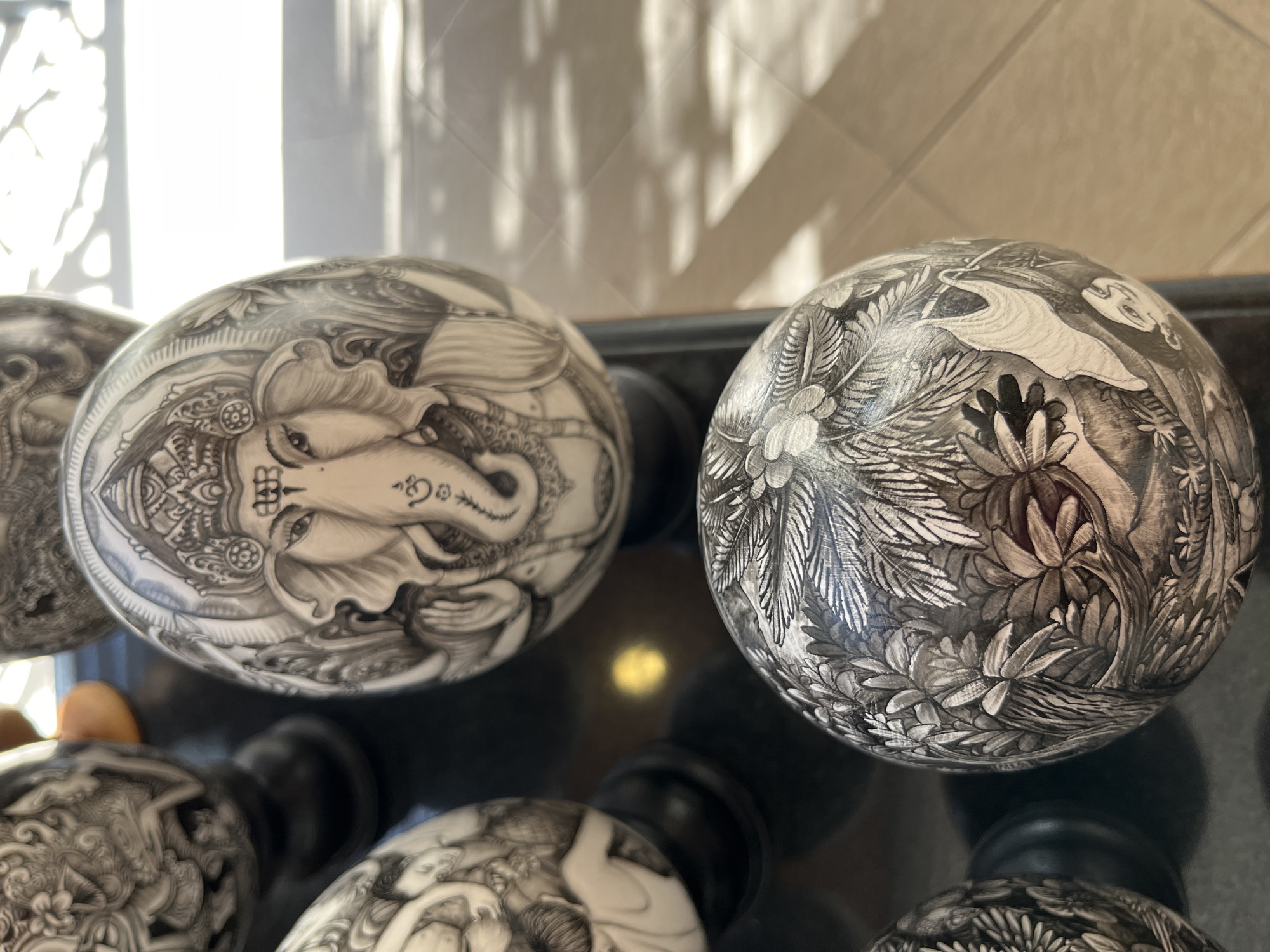 A close-up, angled shot captures three elaborately carved oval objects, possibly ostrich eggs, displayed on a dark, reflective surface. The eggs are intricately detailed with black and white carvings. The foremost object prominently features the head of the Hindu deity Ganesha, adorned with a headdress and jewelry. To its left, another egg shows a depiction of figures in a naturalistic scene. The third visible egg, positioned at the bottom, is covered in detailed carvings of lush foliage and flora, with a subtle hint of a creature or human form integrated into the design. The background is out of focus, showing a tiled floor with dappled sunlight creating a geometric pattern of light and shadow, suggesting an indoor setting with natural light filtering in, potentially in a shop or gallery in Kuta Selatan, Indonesia.