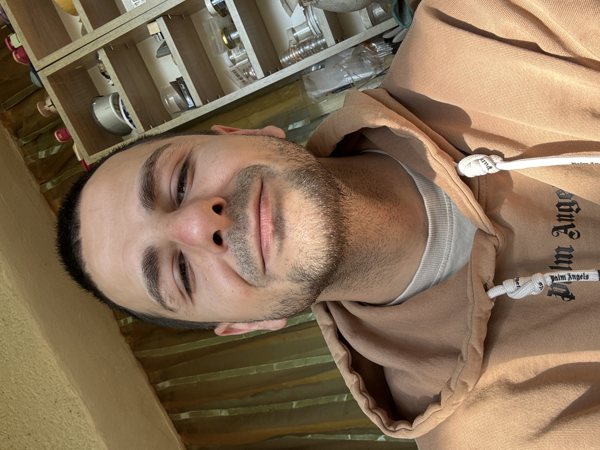 A selfie of a light-skinned man with short dark hair and a beard, wearing a tan hoodie with black lettering, is captured indoors. The background features a shelf with various small objects, and the setting appears to be Slavyani, Bulgaria. The man is positioned to the left, with his face angled slightly toward the camera.