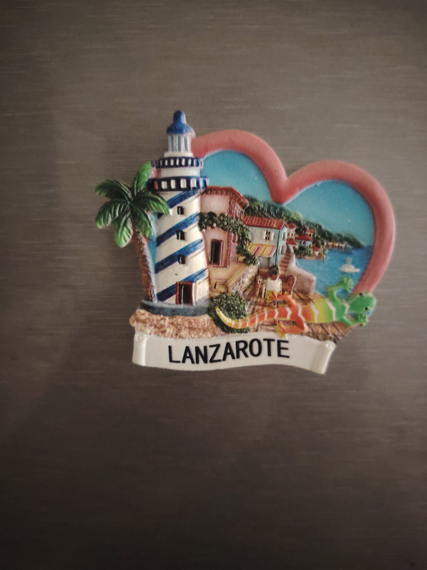 The image displays a colorful, three-dimensional souvenir magnet resting on a dark, horizontally textured surface. The magnet features a heart-shaped outline with a pale red border enclosing a vibrant blue sky and green landscape. Within this scene, a white lighthouse with prominent blue helical stripes and a blue top stands on the left. Adjacent to the lighthouse is a green palm tree with a brown trunk. To the right of the lighthouse, a village with various small buildings, featuring red roofs and light-colored walls, is depicted cascading down towards a blue body of water, possibly a bay, where a small white sailboat is visible. In the foreground, below the village, a prominent, stylized lizard with an orange, yellow, and green gradient body lies on the rocky terrain. A white banner at the bottom of the magnet features the word "LANZAROTE" in black capital letters, identifying the depicted location as Lanzarote, an island in the Canary Islands.
