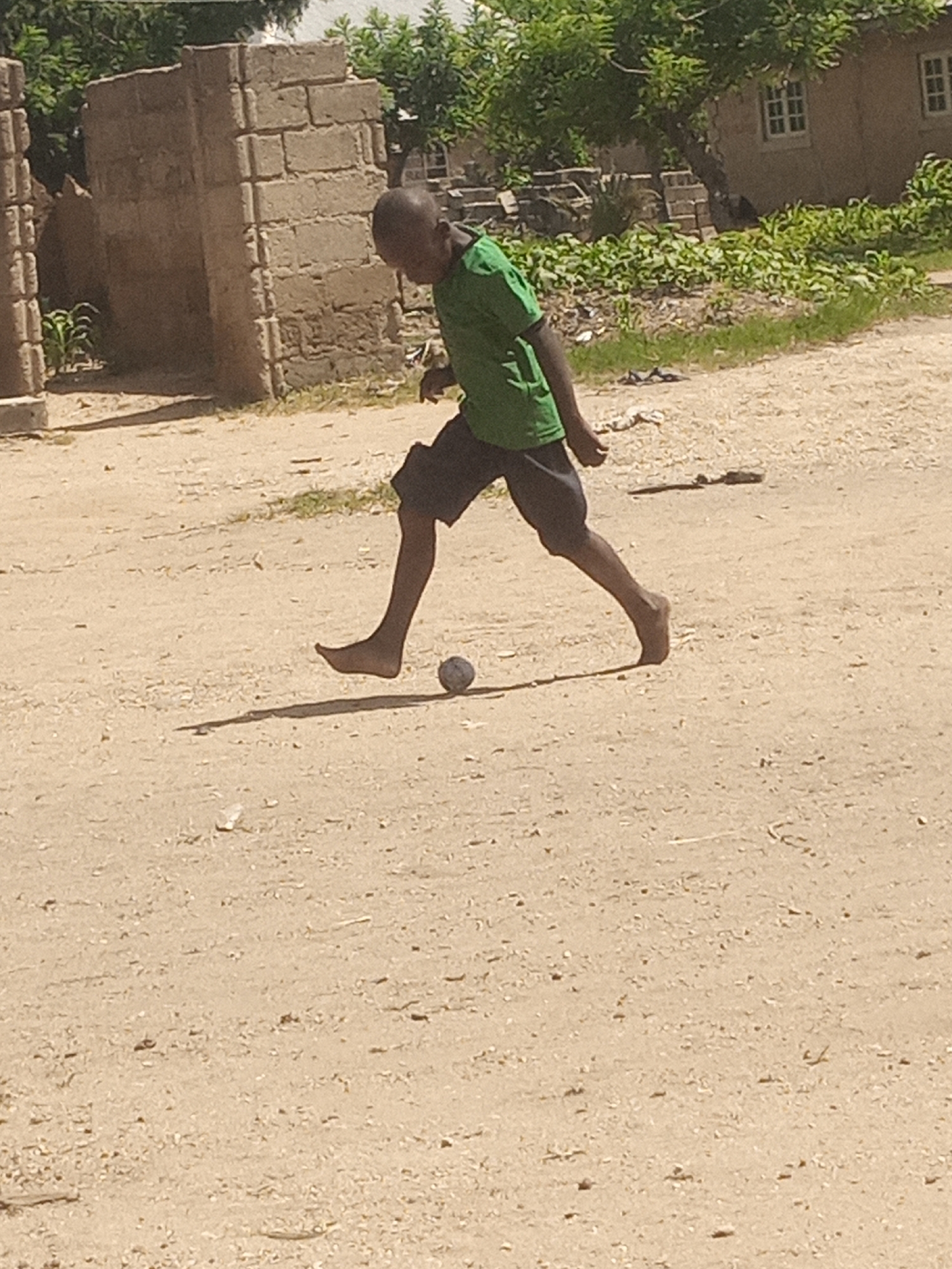 A young boy, appearing to be of African descent, is captured mid-stride, barefoot, in what seems to be a casual game of football on a dusty, unpaved surface. He is wearing a green t-shirt and dark shorts. His body is angled as if kicking or controlling a small, greyish ball at his feet. The environment suggests a rural or semi-urban setting in Langtang, Nigeria, characterized by rudimentary brick structures with visible mortar, overgrown vegetation, and scattered debris on the ground. The lighting indicates a sunny day, casting distinct shadows. In the background, another brick building with windows and foliage can be seen, adding to the sense of a developing or undeveloped area.
