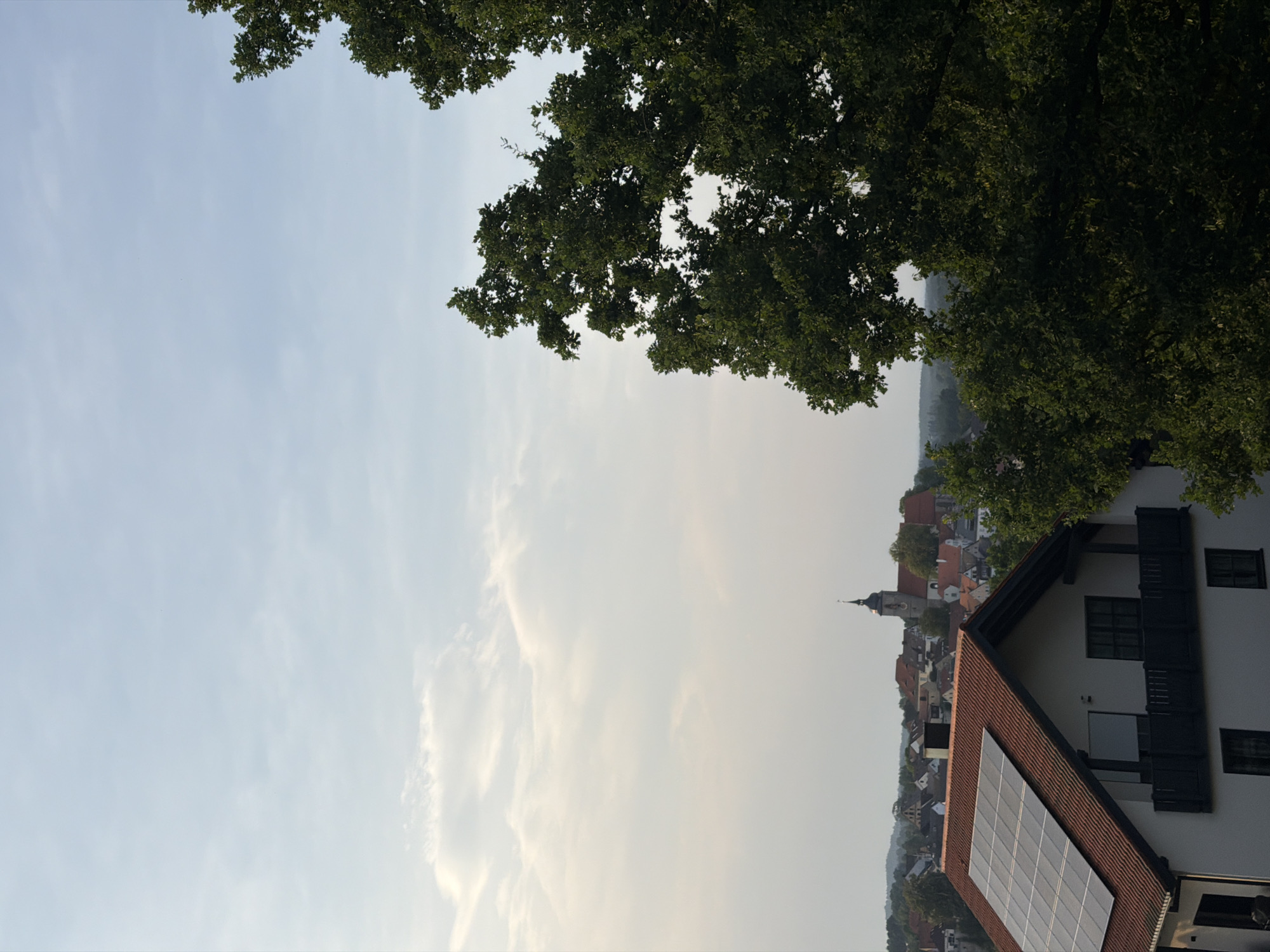 The image shows a rural landscape with a town in the background, likely Creußen, Germany. In the foreground, a white house with a red tiled roof is visible, equipped with solar panels. A large tree with lush green foliage dominates the right side of the frame. In the distance, a church spire with a dark roof rises above a cluster of buildings with red roofs, characteristic of a German town. The sky is partly cloudy with a light, hazy appearance. No people or animals are visible in the image.
