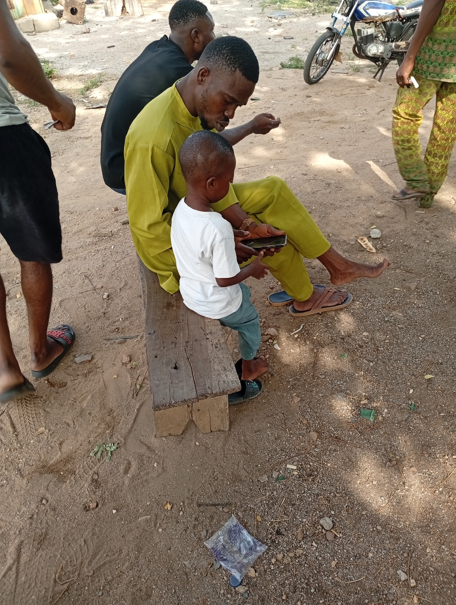 The scene is located in Adankolo, Nigeria. Three adult men and a young boy are present outdoors on a sandy, dirt-covered ground. Two men are seated on a weathered wooden bench. The boy is seated on the end of the bench next to one of the seated men, and both are looking at and interacting with a mobile phone held by the man. The seated man wears a yellow shirt and pants and brown sandals. The boy is wearing a white shirt and light blue pants and black shoes. Another man wearing a black shirt is seated on the bench beside the man in yellow. A fourth person is partially visible to the left wearing a grey shirt and black shorts and black shoes. A fifth person is also partially visible on the right, wearing green patterned pants. A motorcycle is parked in the background. A plastic bag is on the ground near the bench.