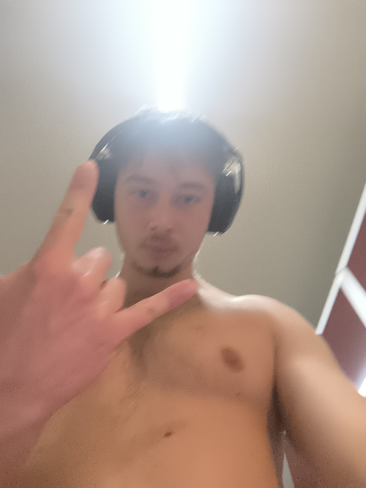 A shirtless young man with headphones stands indoors, holding up the "rock on" hand gesture. He has light skin, short dark hair, and a mustache. The background is blurry, but there is a light-colored wall and a red-and-white door or panel. There are no identifiable landmarks that would place the location in Sofia, Bulgaria.