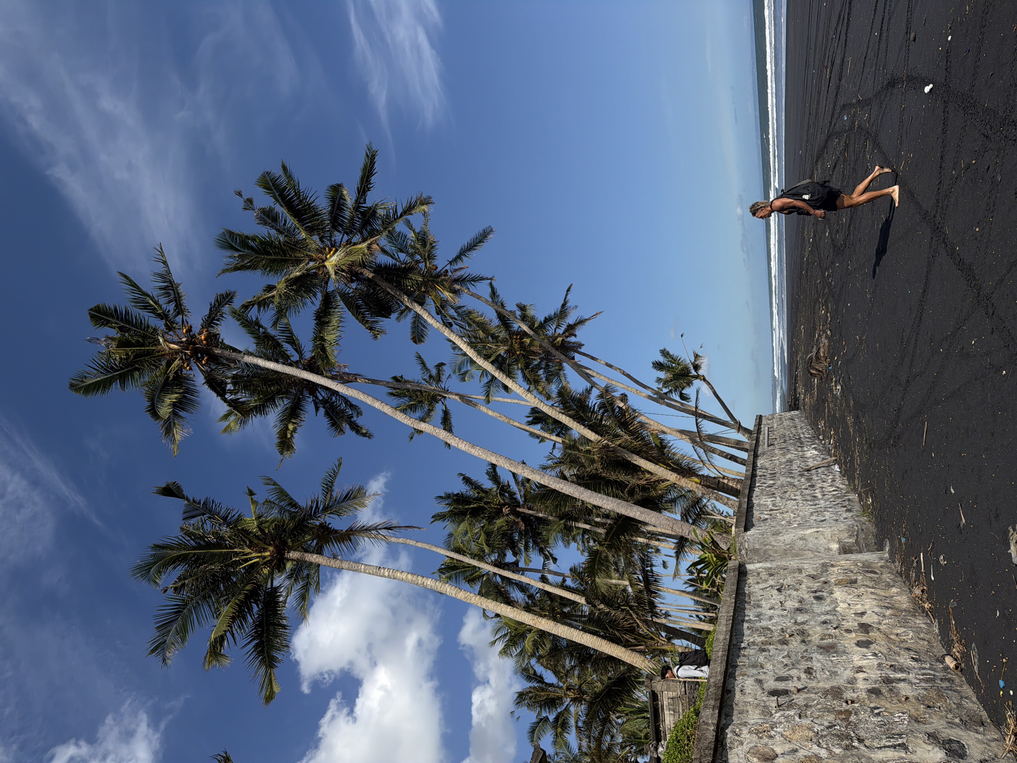 The image shows a beach scene in Kerambitan, Indonesia. Palm trees line a stone wall that separates the beach from land. A man wearing dark clothing walks barefoot on the black sand beach toward the ocean. The sky is blue with scattered clouds. The ocean is visible in the background, with small waves breaking on the shore.