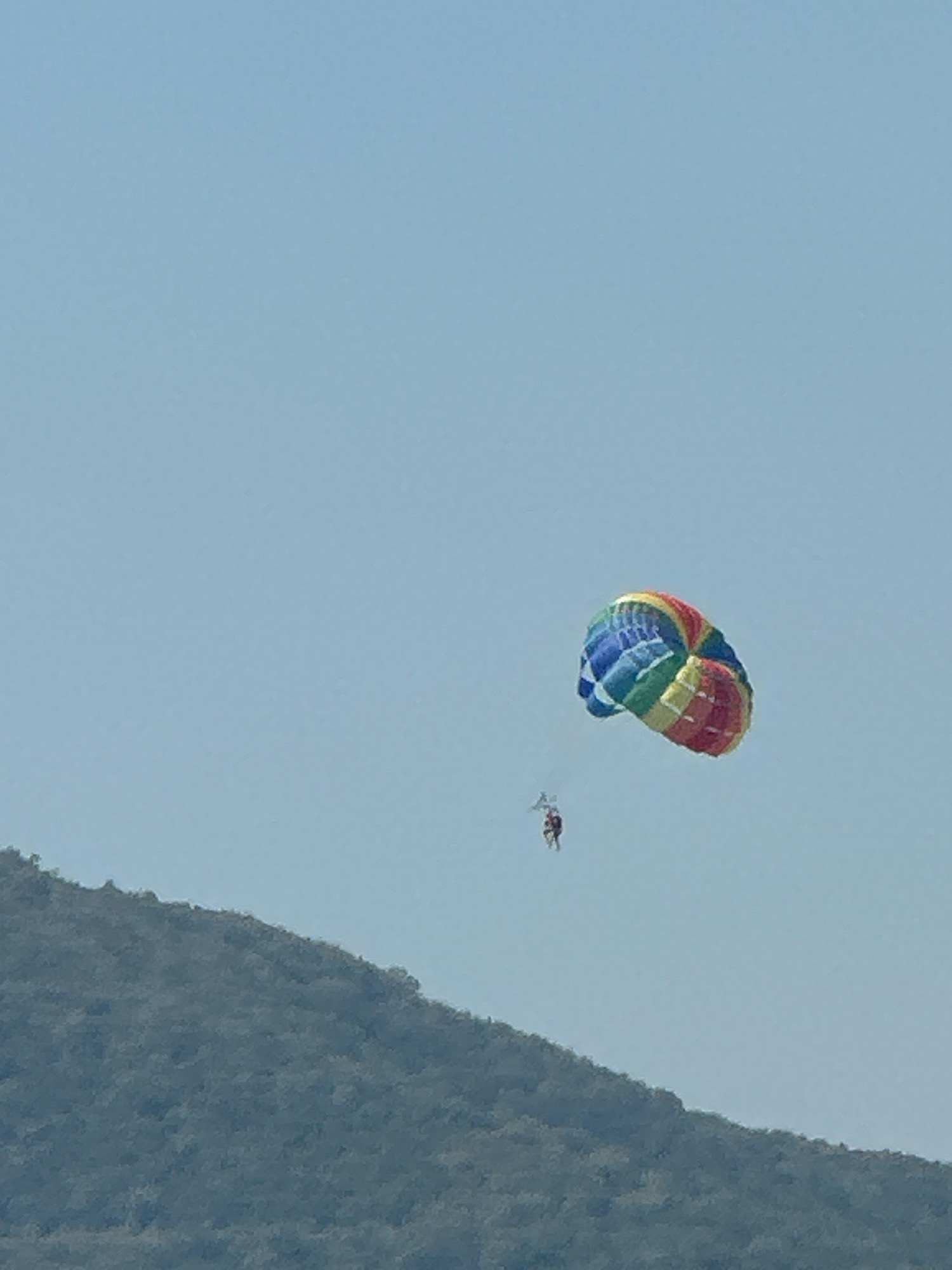 A person is parasailing against a clear blue sky with a lush green mountain range in the foreground. The parasail is a vibrant rainbow of colors, and the person appears to be suspended in mid-air, enjoying the activity. The image captures a moment of adventure and recreation in a picturesque natural setting. The location is the coast of Budva, Montenegro, a popular tourist destination known for its beautiful beaches and stunning scenery. The parasailer is a single individual.