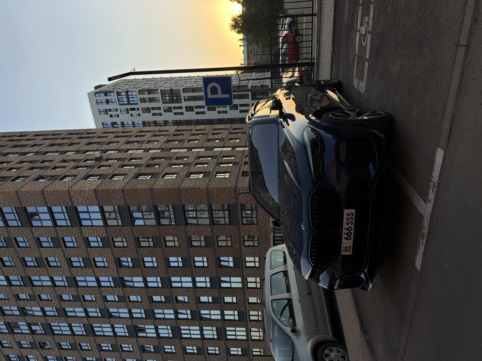 The image depicts a street scene in Izhevsk, Russia, near modern residential buildings. A dark blue BMW SUV with license plate "04 666 SSS" is parked prominently. A light-colored vehicle is parked behind the BMW. A parking sign with a "P" is visible near the front of the BMW. The backdrop features modern apartment buildings with brick and white facades. The time of day appears to be near sunset, with the sun visible in the upper part of the frame. The overall environment is urban, with clear parking markings on the asphalt surface. A metal barrier separates the parked cars from a sidewalk.